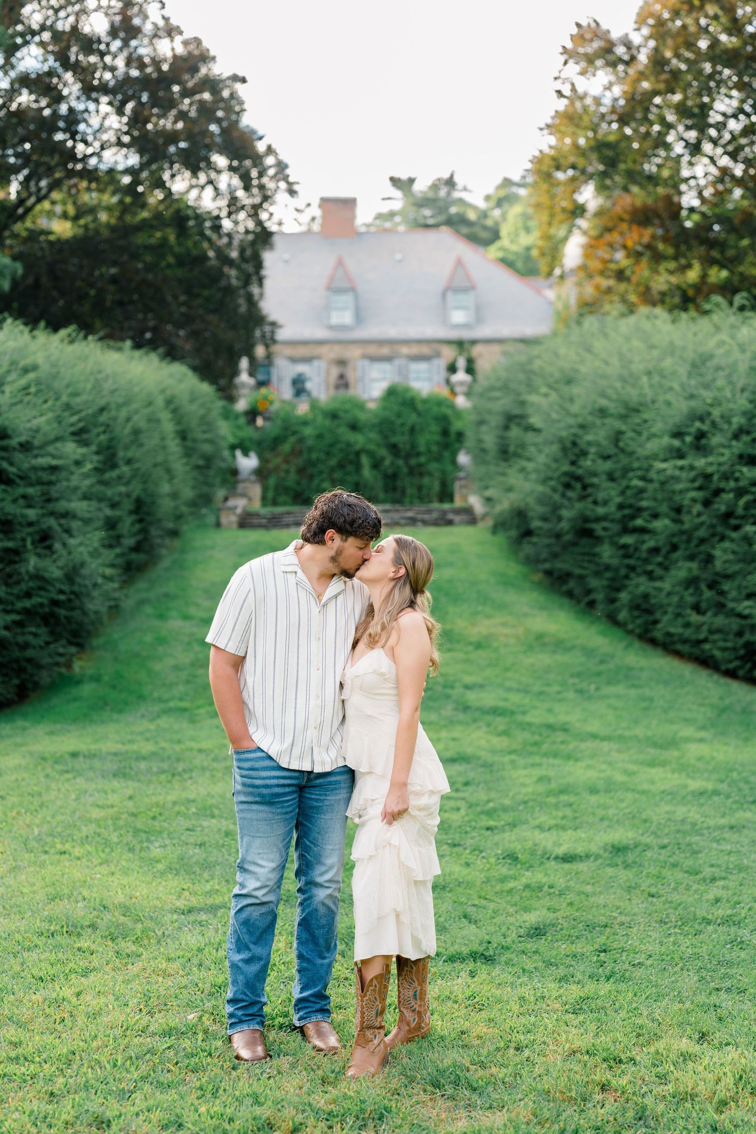 Couple kissing in lush garden pathway with historic manor house backdrop, woman in white ruffled dress and cowboy boots.