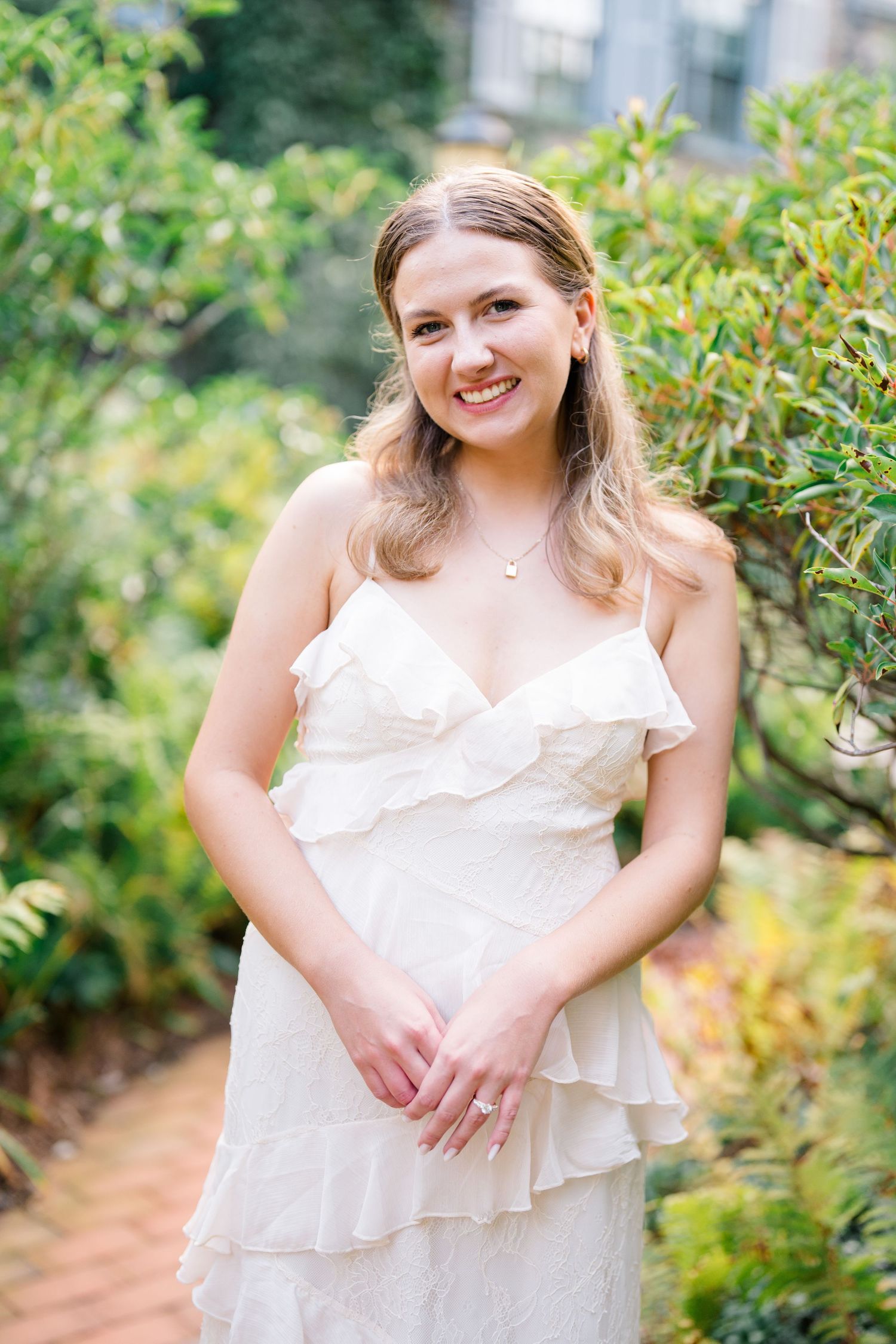 Smiling young woman in white ruffled dress posing in lush green garden setting.