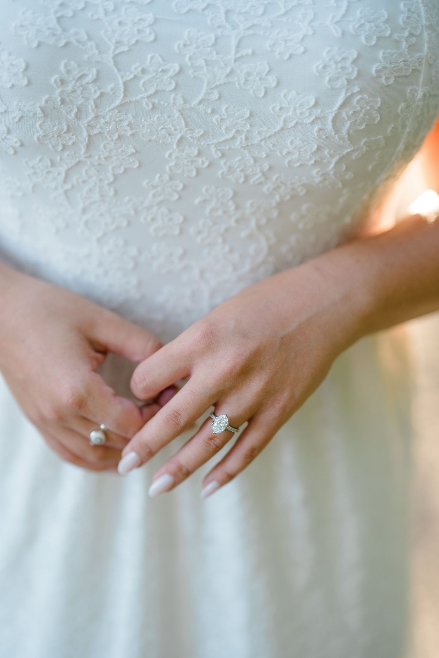 Close-up of bride's hands showing elegant diamond engagement ring and pearl ring against lace wedding gown.