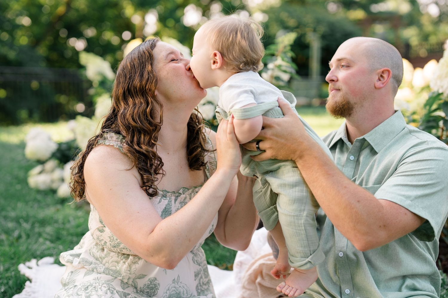 Happy family outdoors, mother kissing baby held by bald father in green shirt, lush garden background.