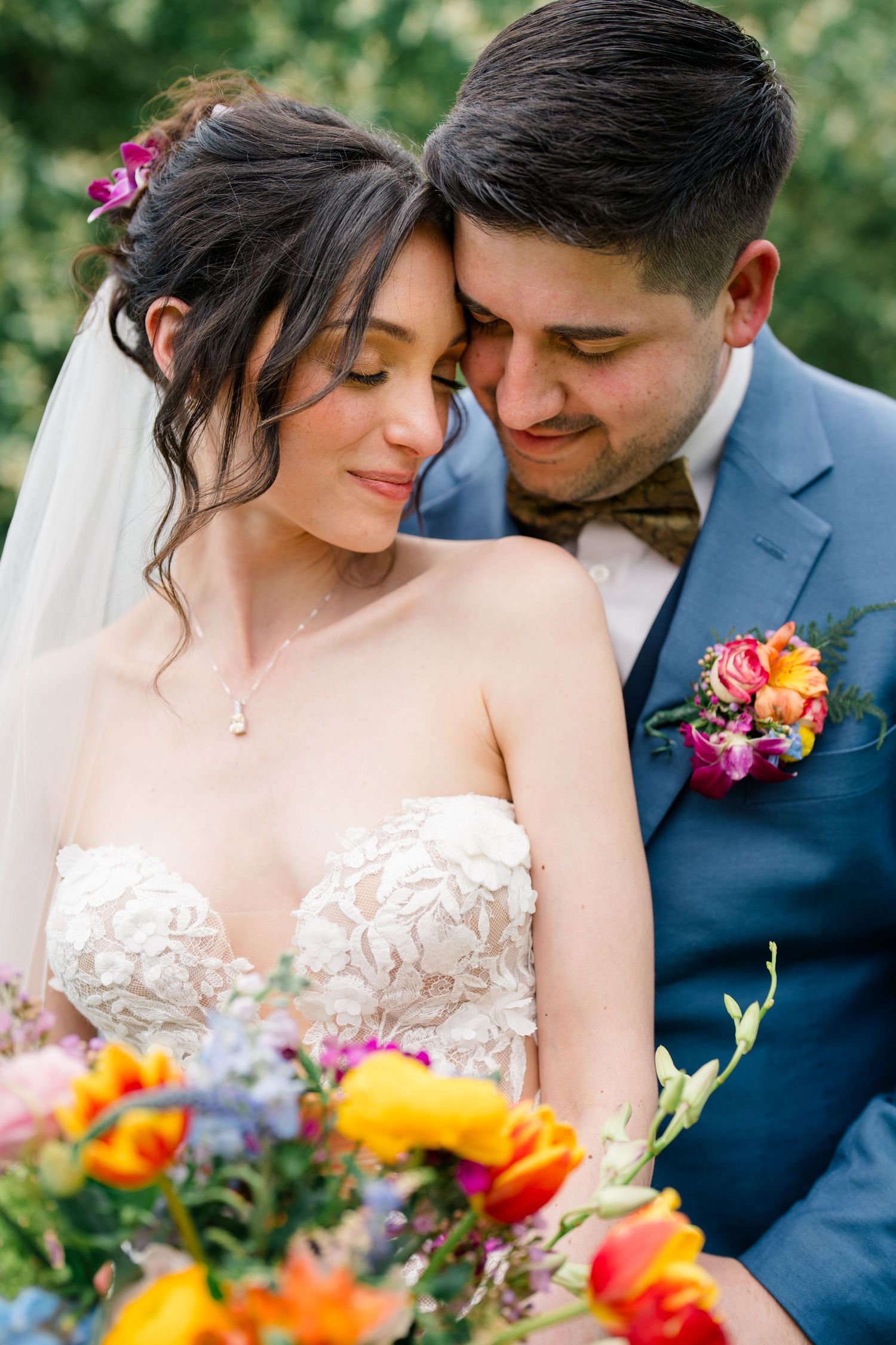 A bride in a delicate lace gown and groom in navy suit share an intimate moment surrounded by vibrant flowers.