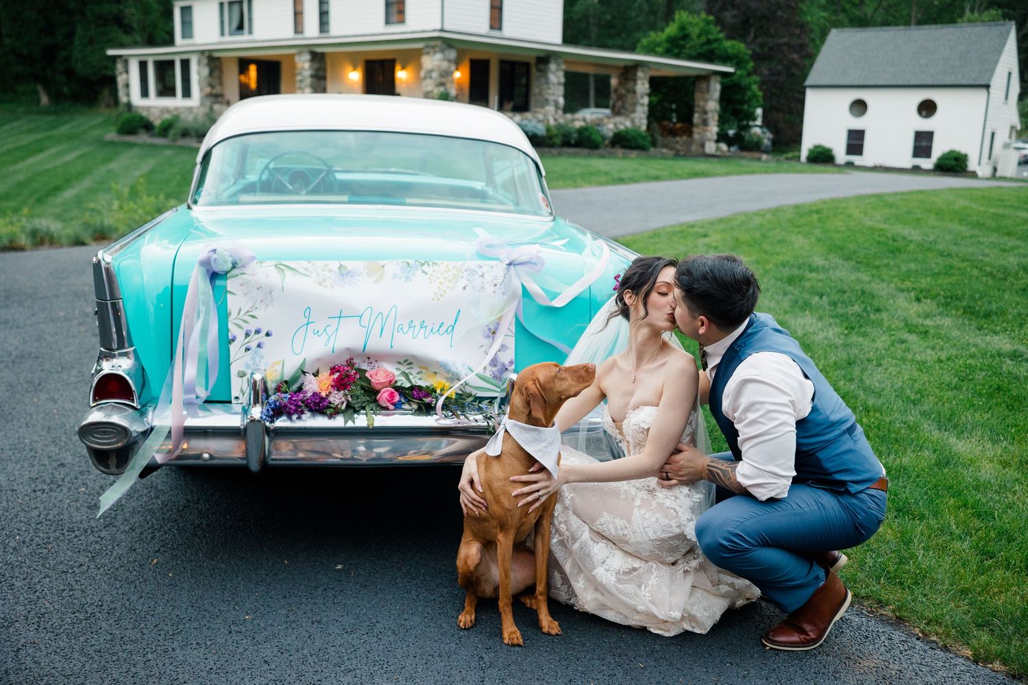 A vintage turquoise Chevrolet with 'Just Married' decorations parked outside a white farmhouse with couple and dog.