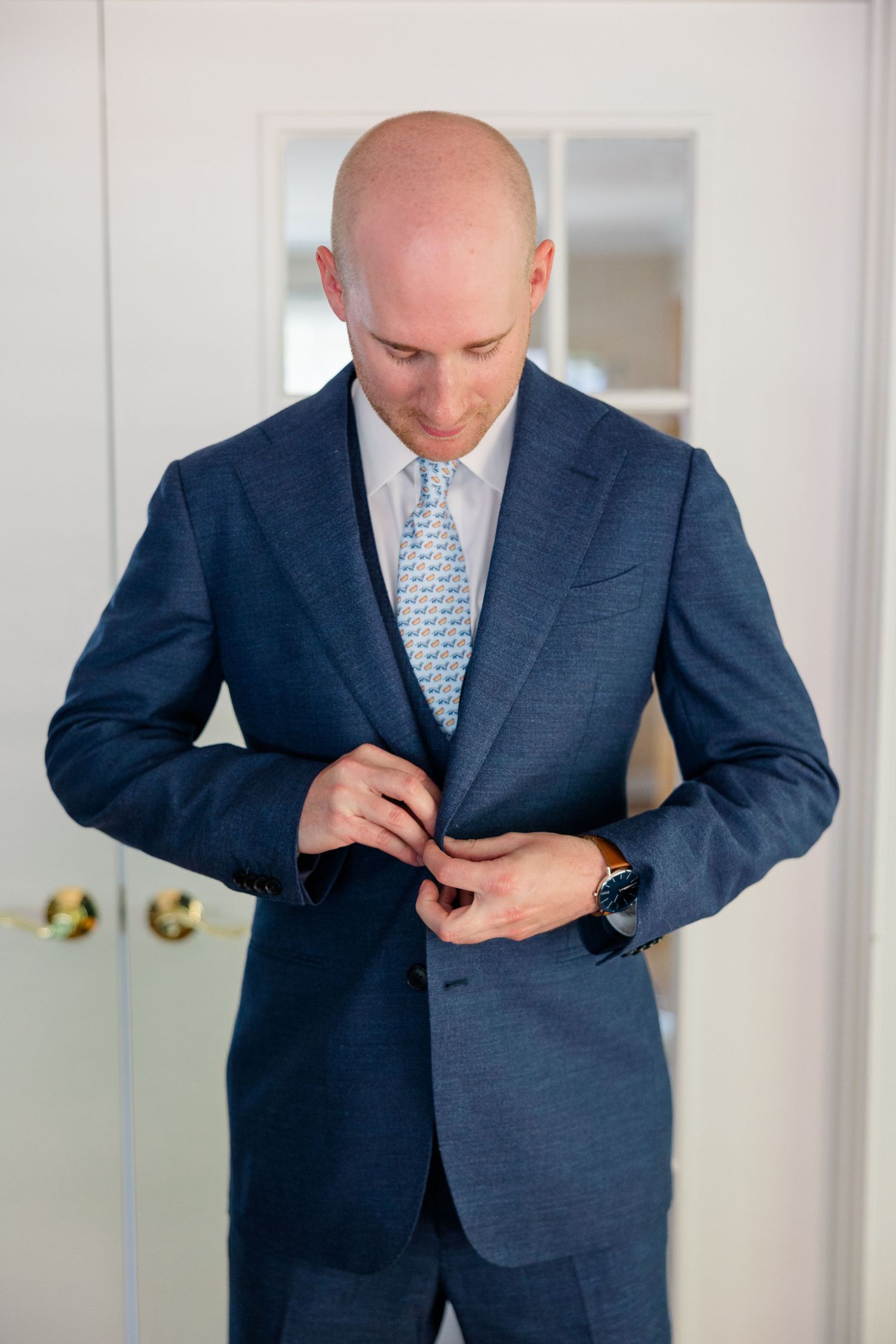 Bald groom in navy blue suit buttoning jacket, wearing light blue tie and watch, standing by white door.