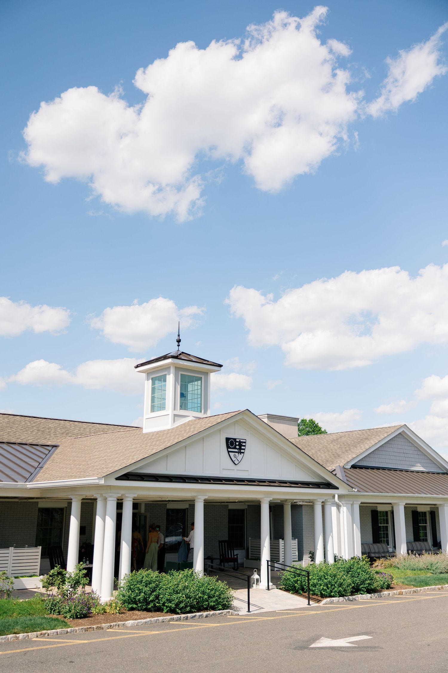 Elegant country club clubhouse with white columns and cupola under a bright blue sky with fluffy clouds.