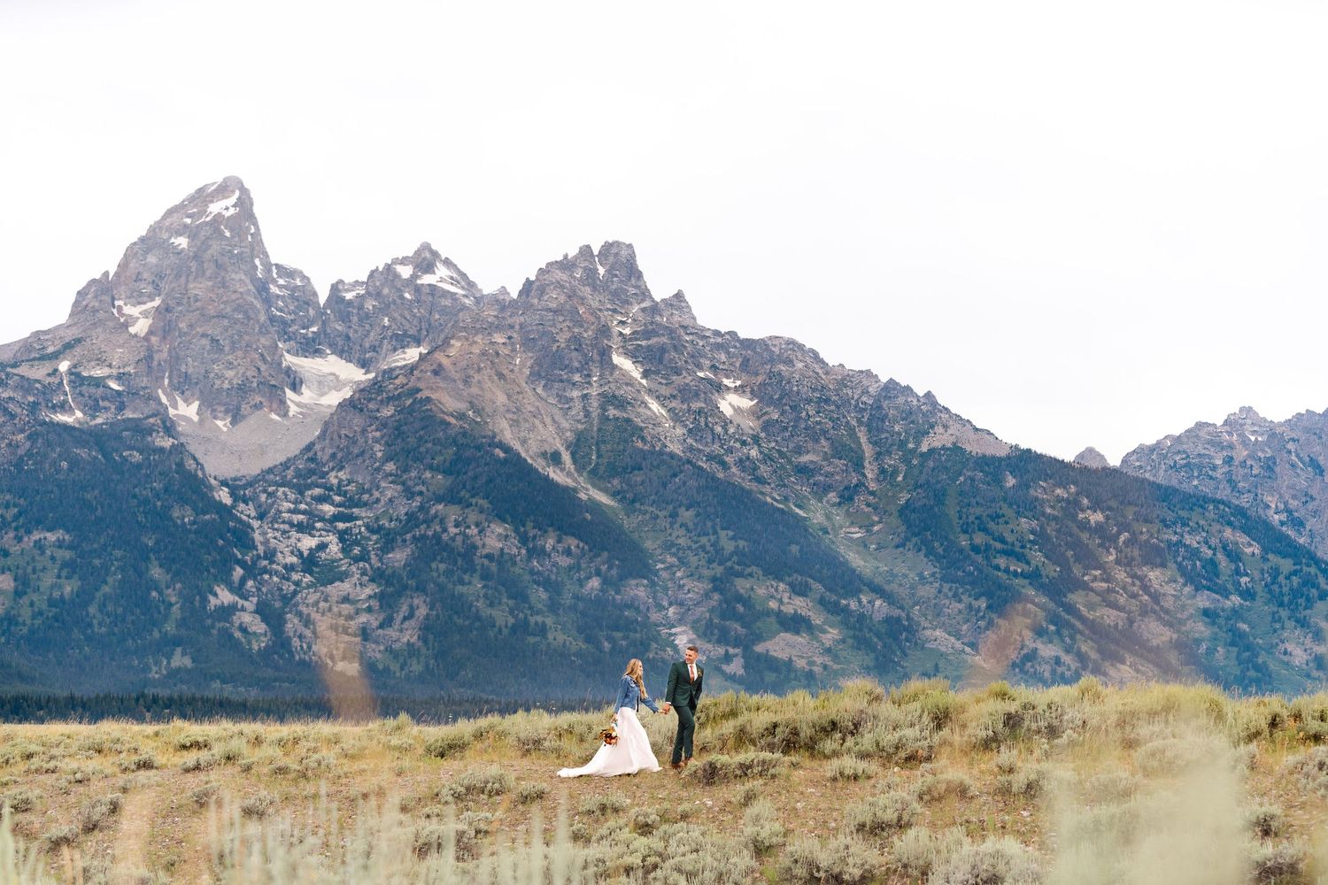 Bride and groom stand in a vast meadow with the dramatic Grand Teton mountain range as a stunning backdrop.