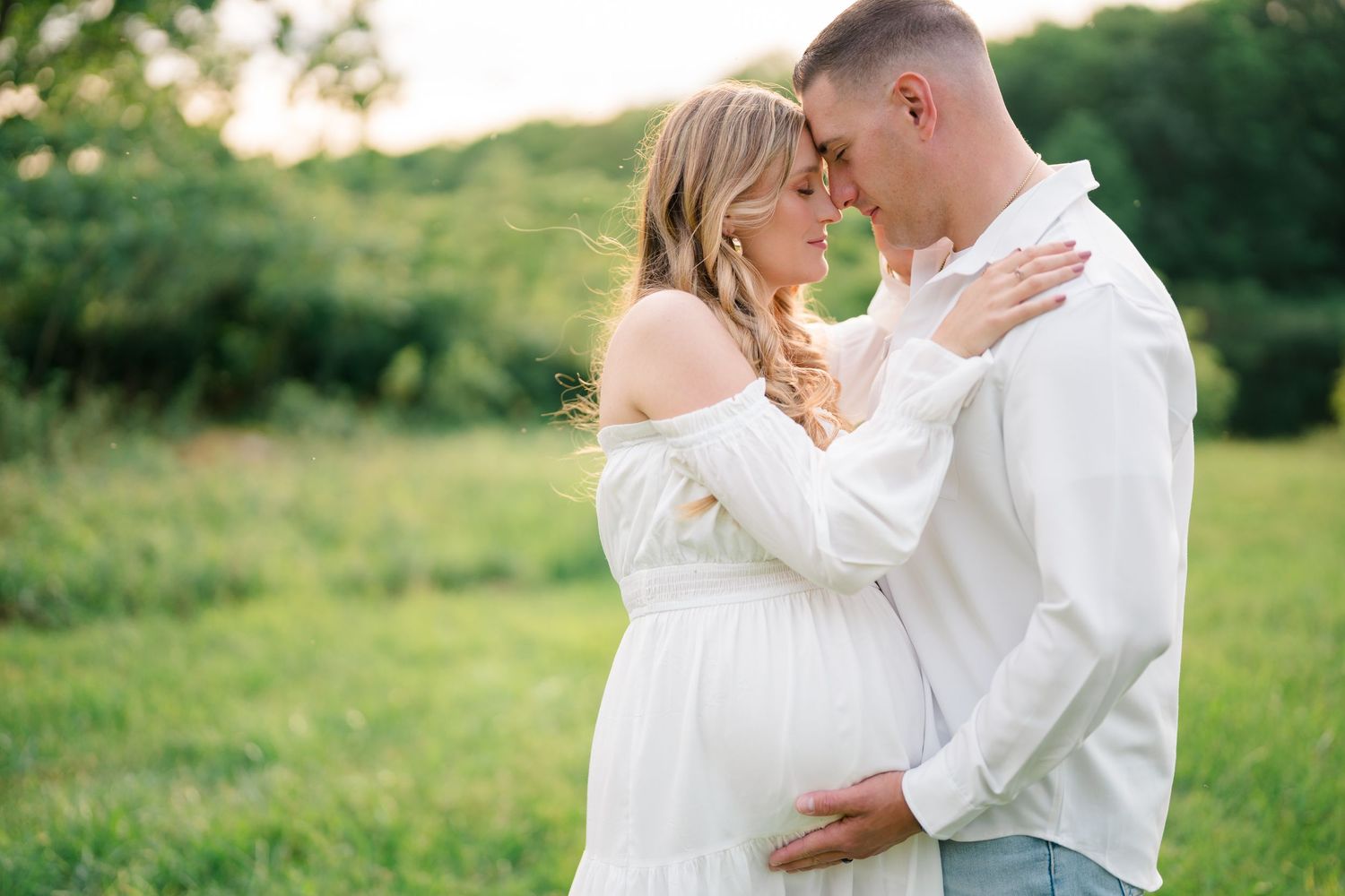 Tender maternity moment captured in outdoor setting with flowing white dress and natural backdrop.