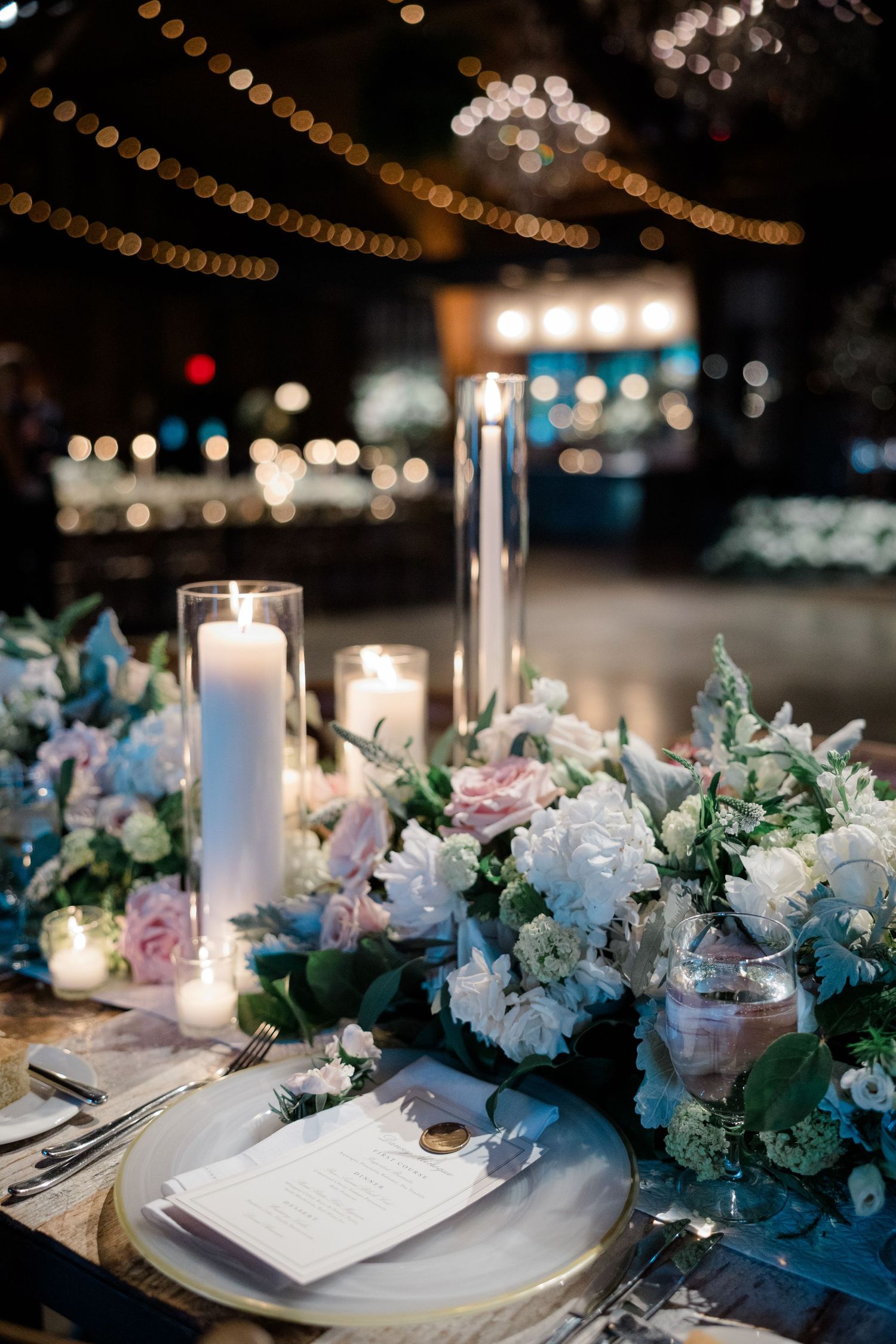 Romantic table setting with candles, blue and pink flowers, and string lights overhead.