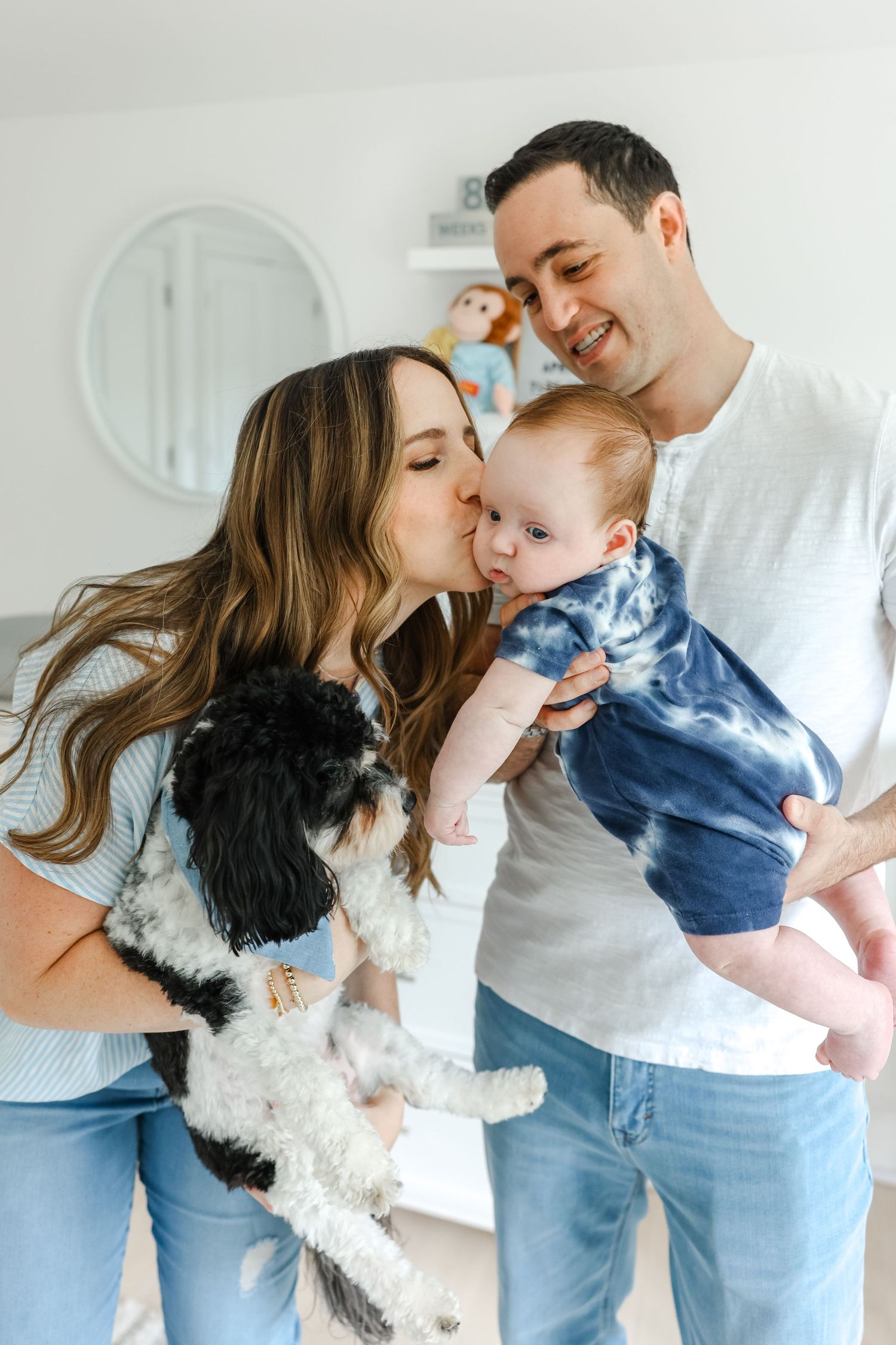 A family shares a tender moment with their baby and black and white dog against a white wall background.