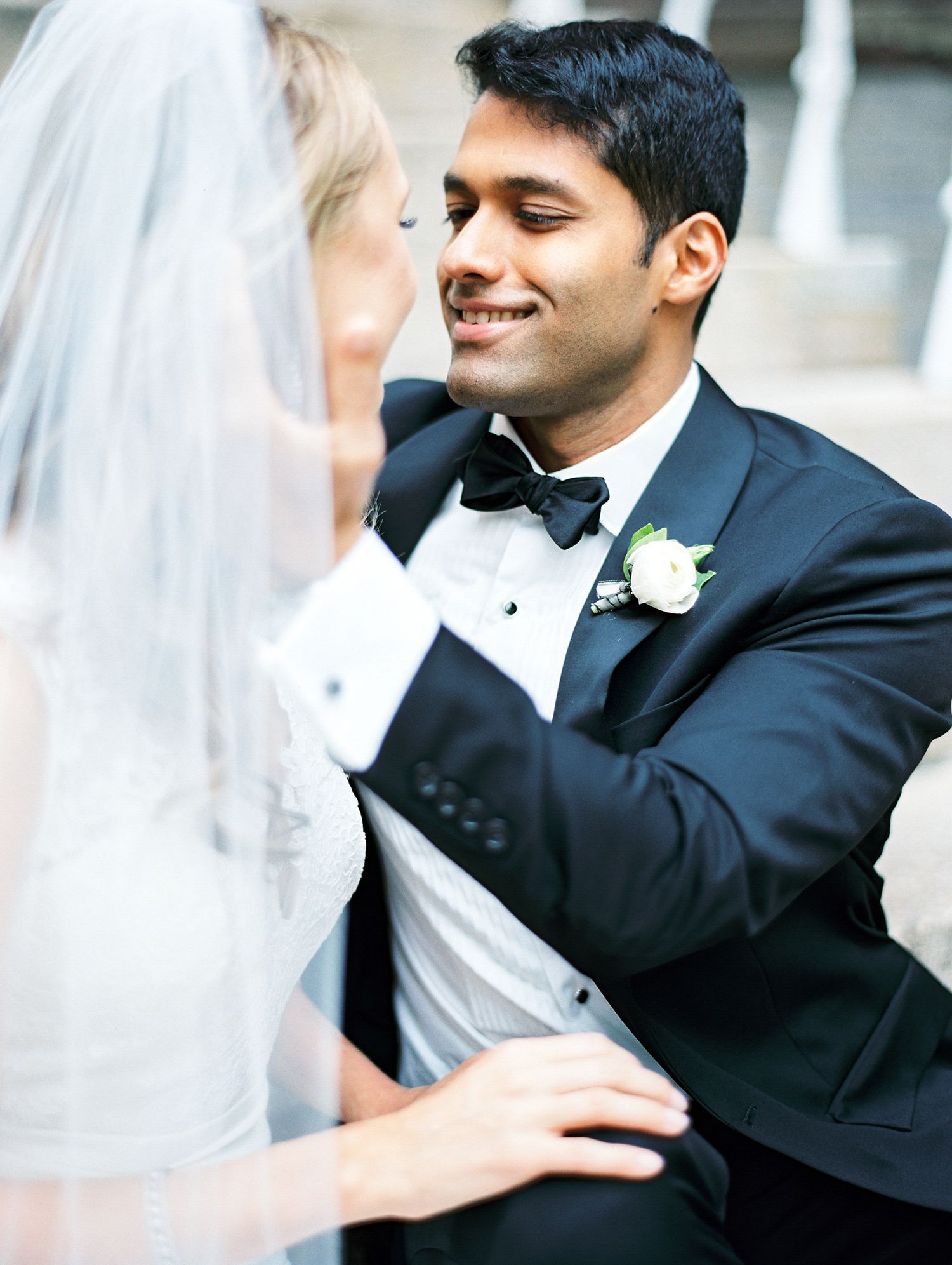 A tender moment between bride and groom on their wedding day, with the groom in a classic black tuxedo and white boutonniere.