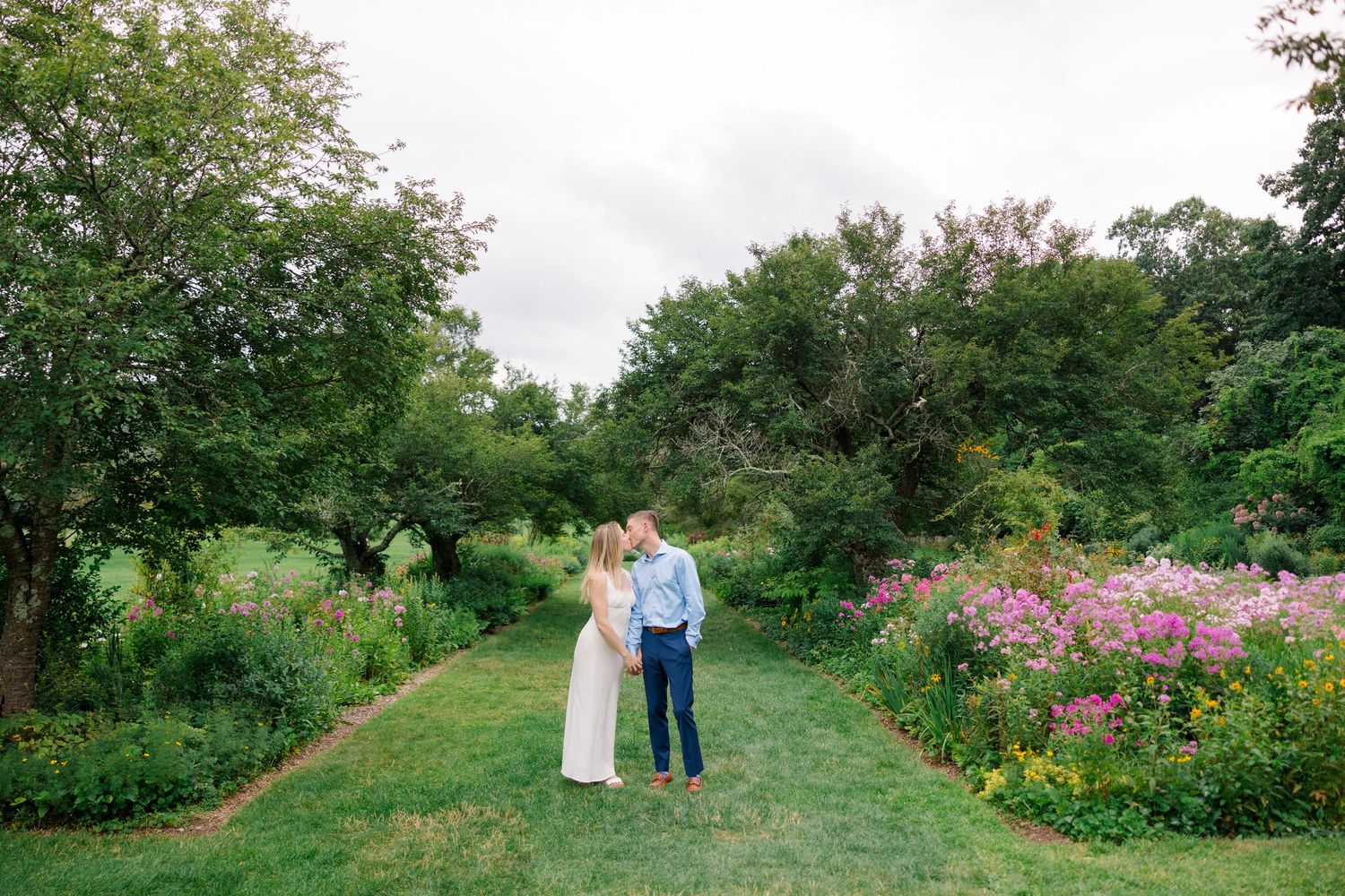 Couple shares romantic kiss in lush garden pathway surrounded by colorful wildflowers and green trees on wedding day.