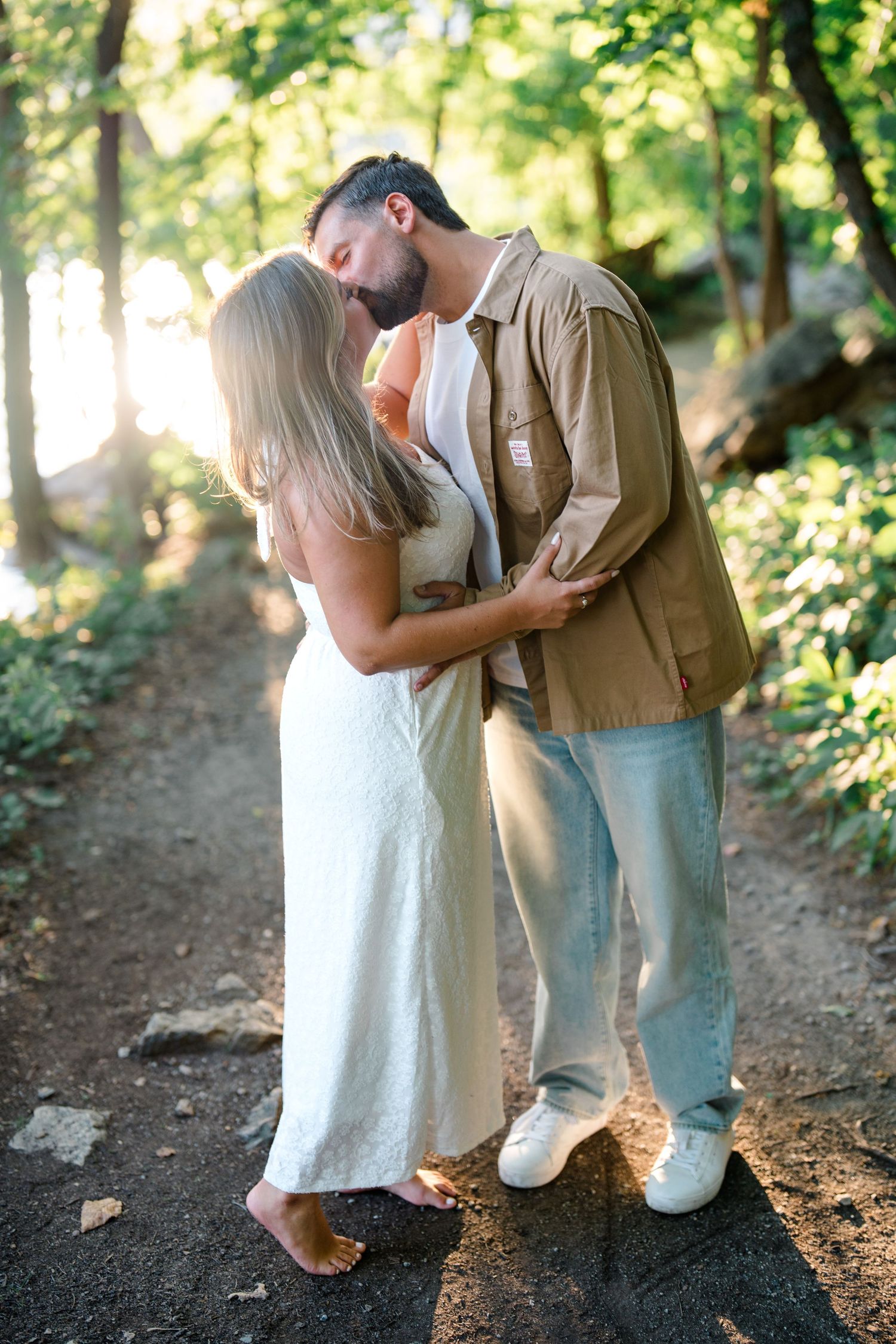 Couple sharing a kiss on a sunlit forest path, woman in white dress, man in khaki jacket and jeans.
