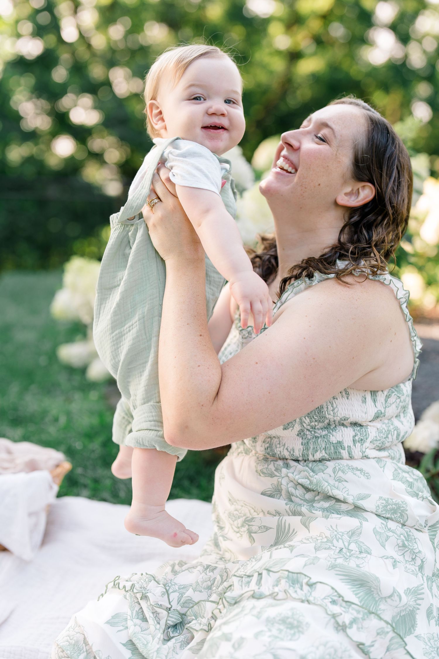 A laughing mother in a floral dress lifts her smiling baby outdoors on a sunny day.