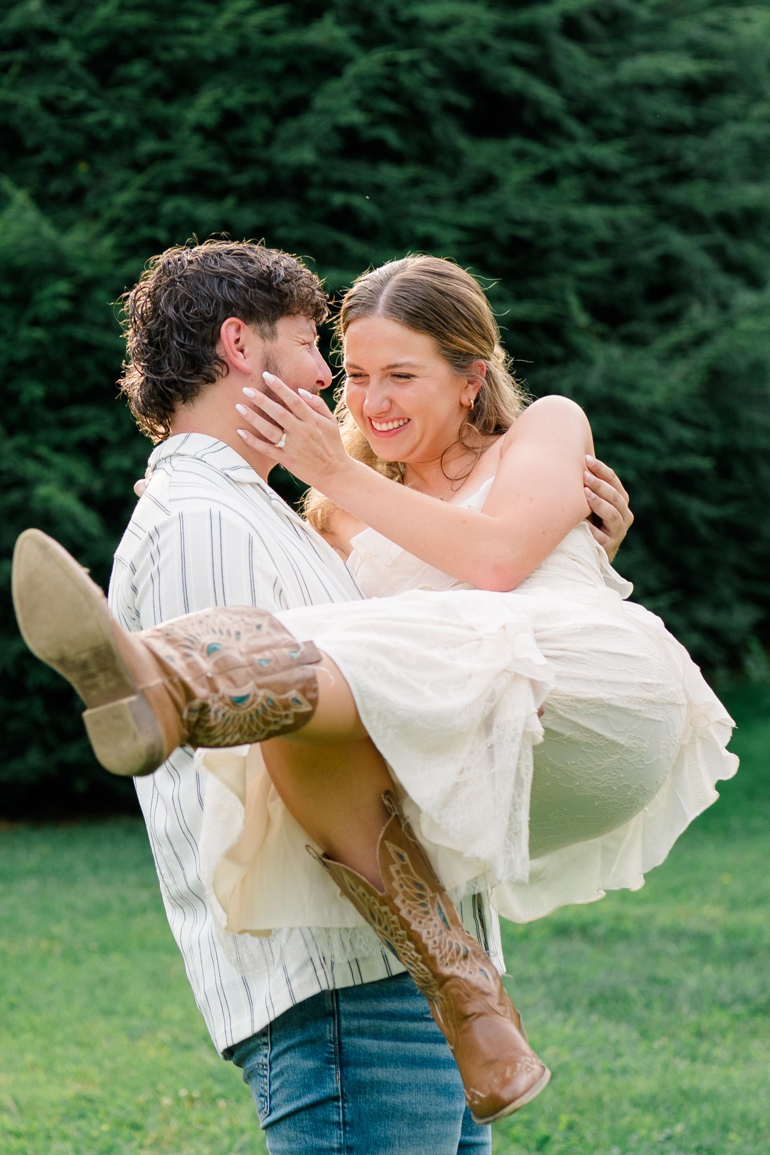 Man with tattoos lifts smiling woman in white dress and cowboy boots outdoors near green trees.
