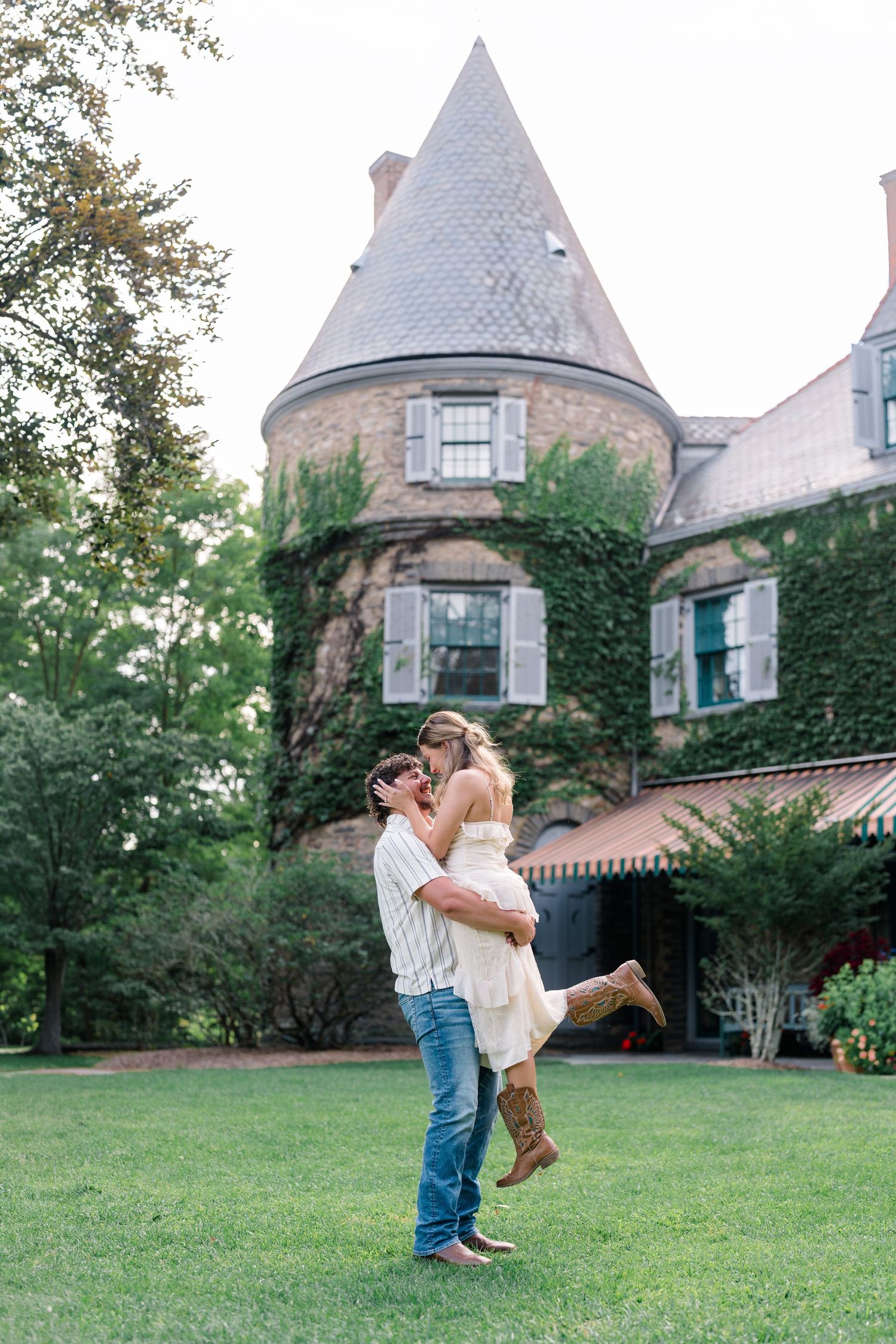 Couple embracing on lawn before ivy-covered stone castle mansion with turret during outdoor engagement photo session.