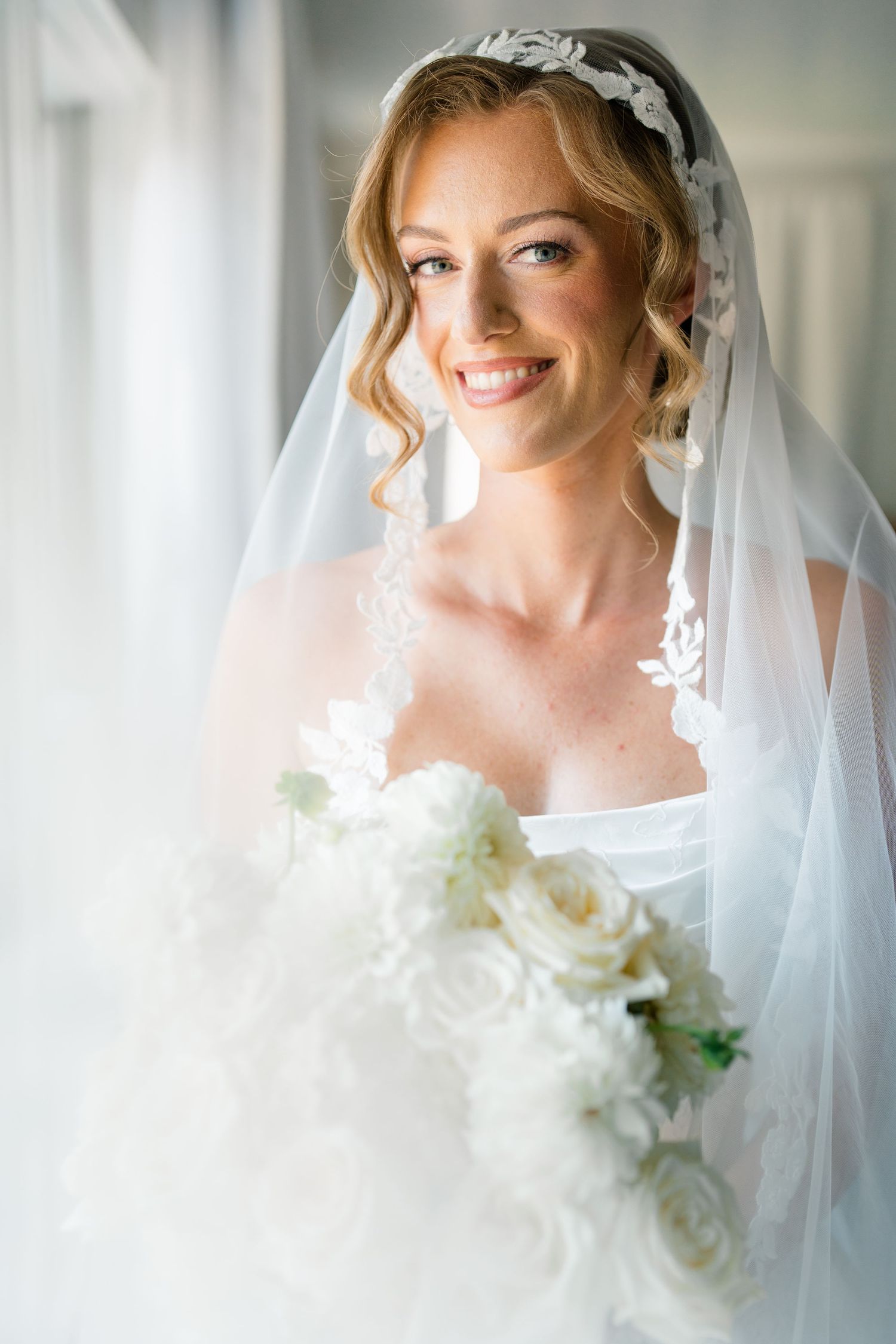 Radiant bride in white gown with lace veil headpiece holding white rose bouquet near bright window.