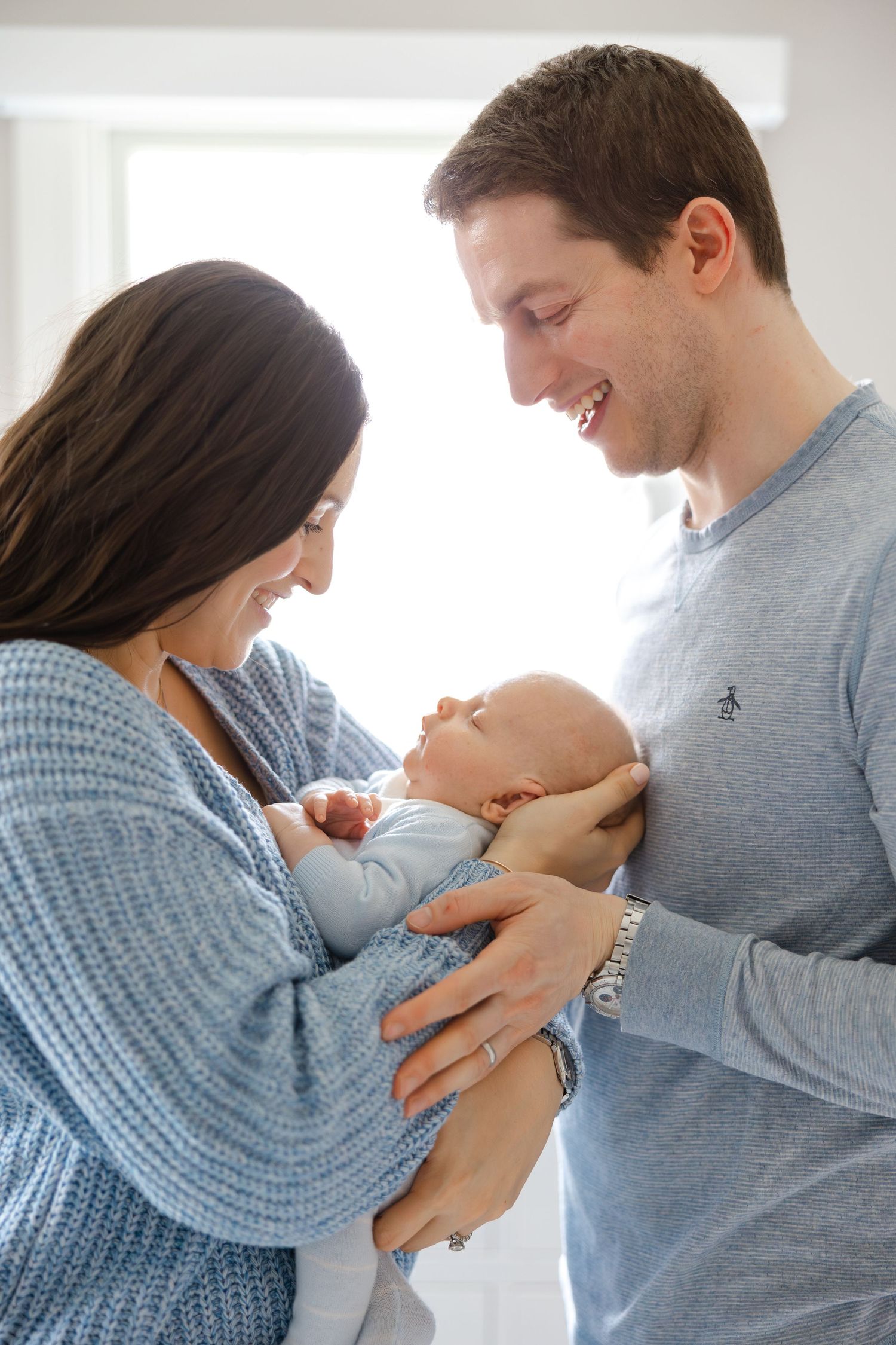 Happy parents in blue sweaters holding and gazing lovingly at their newborn baby in a bright room.