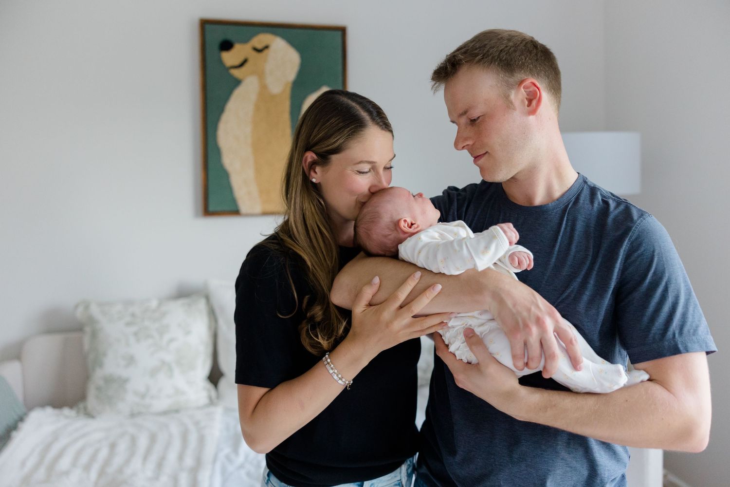 A loving couple in casual attire tenderly holds their newborn baby in a bright white bedroom setting.