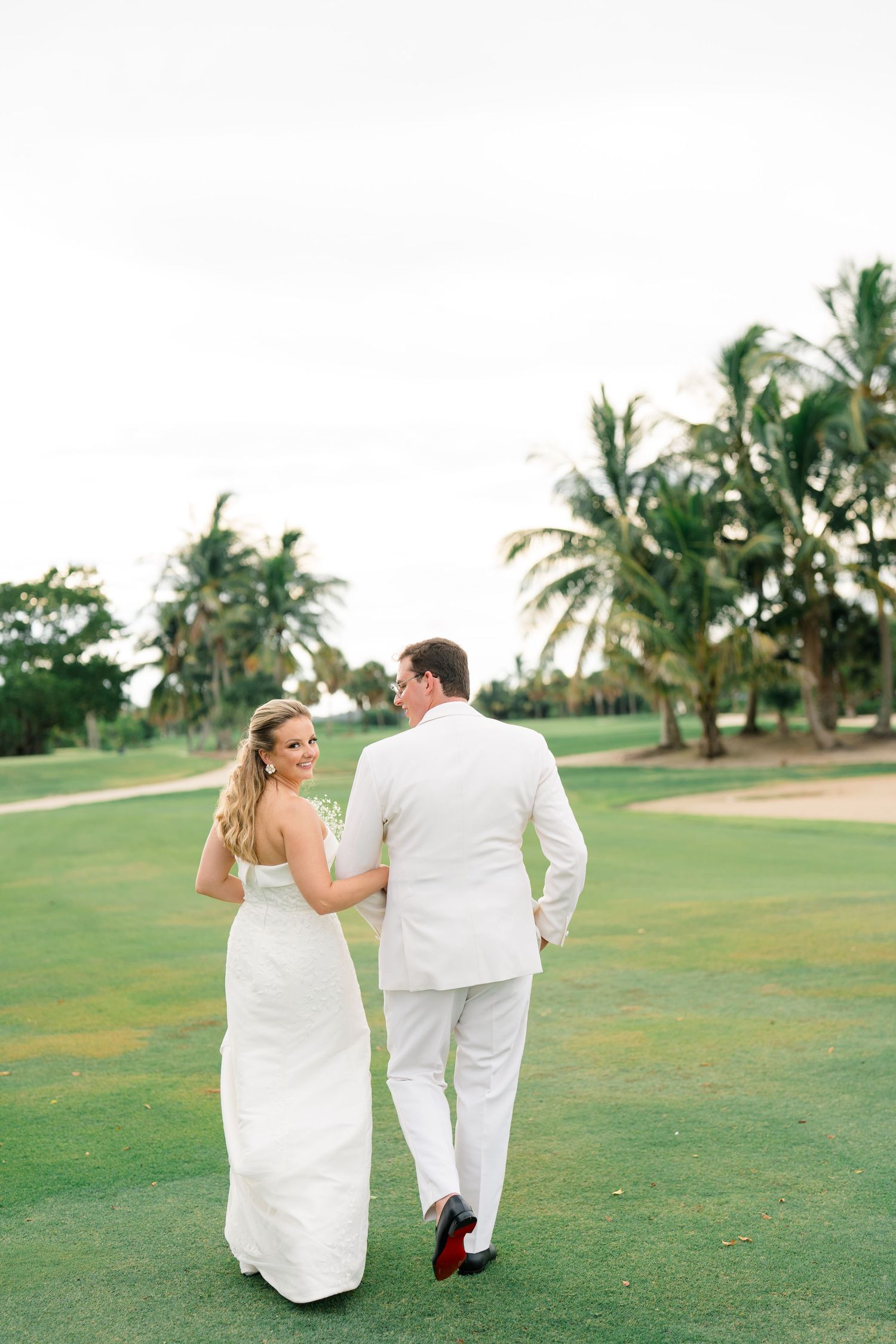 A romantic couple in white attire walks together on a lush green golf course with palm trees in the background.