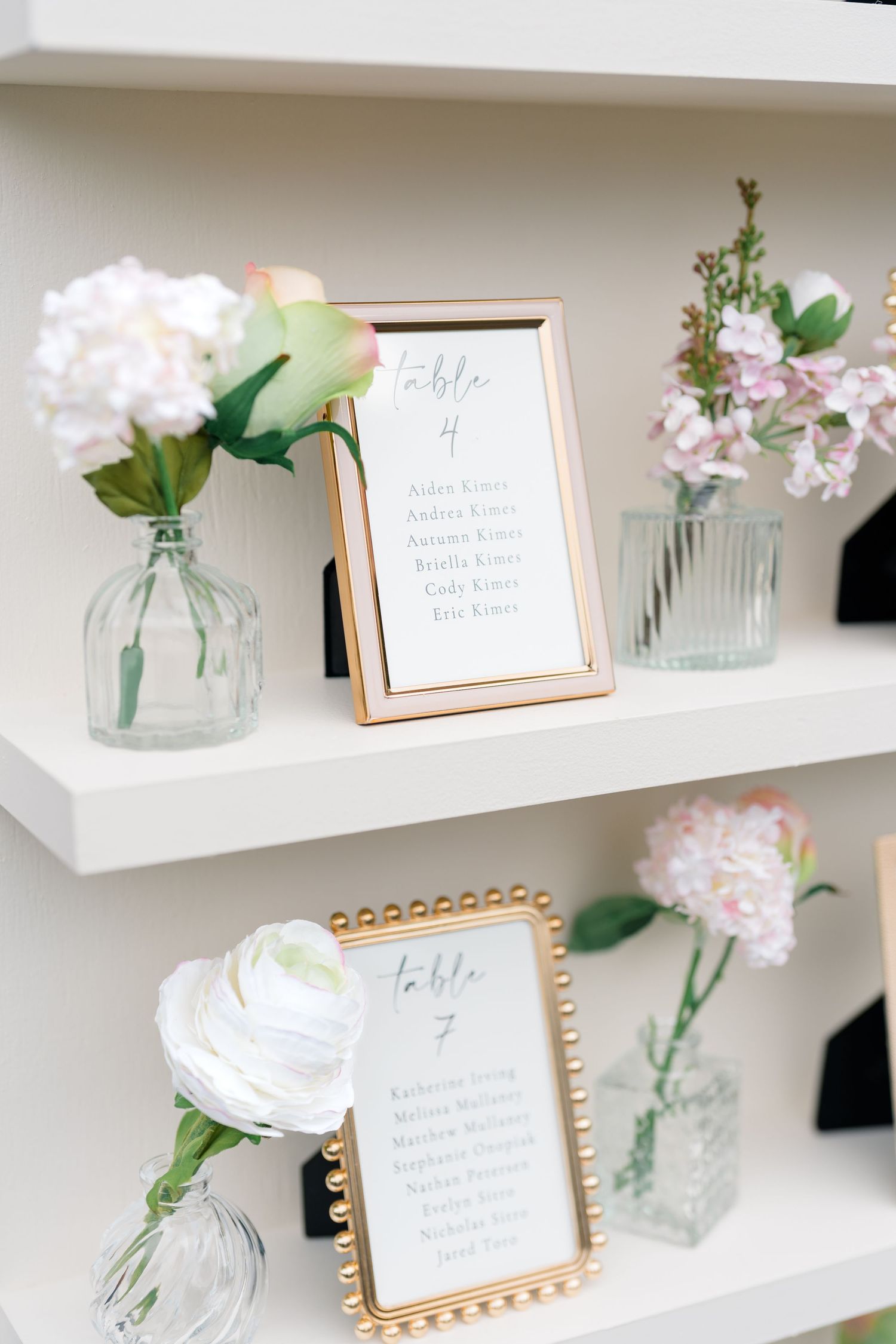 Elegant place card display with floral arrangements and gold frames on white shelves.