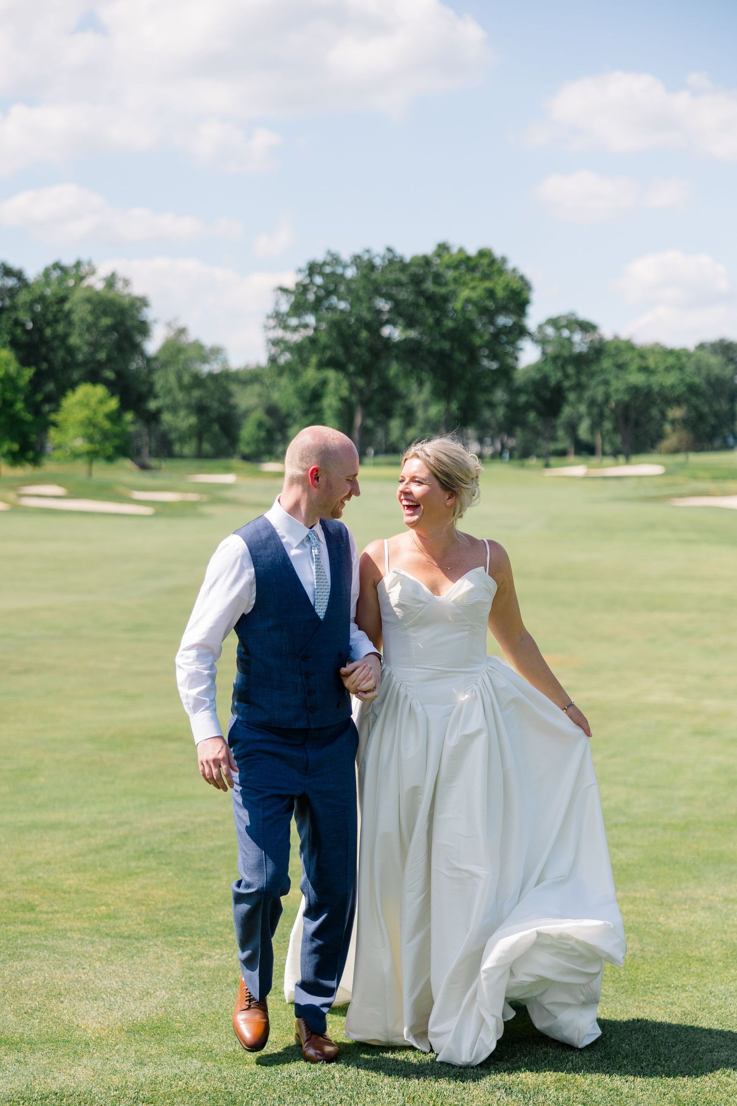 Bride and groom walking hand in hand on a sunny golf course, smiling at each other in wedding attire.