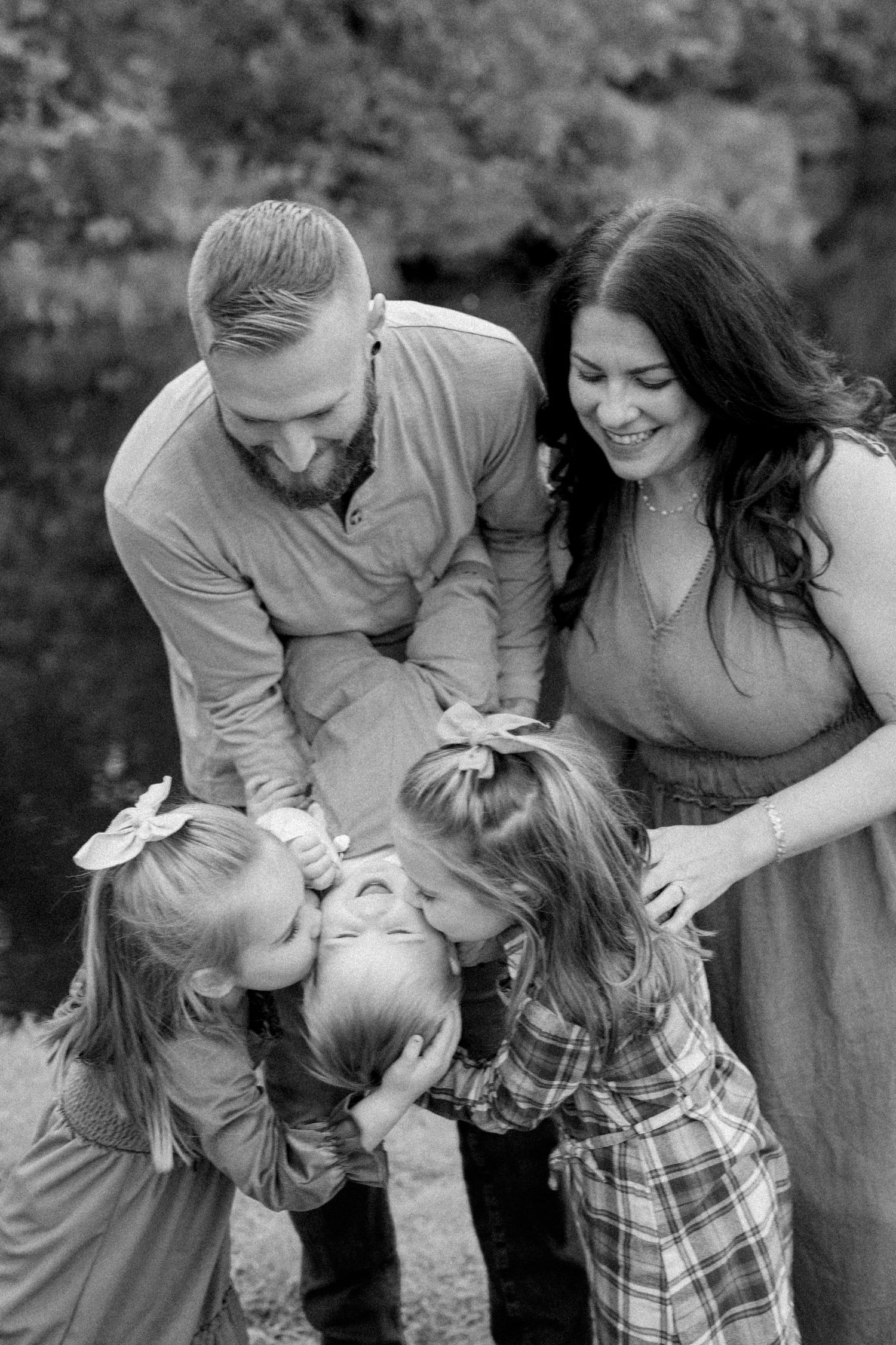 Black and white family portrait of parents with three young children sharing kisses outdoors.