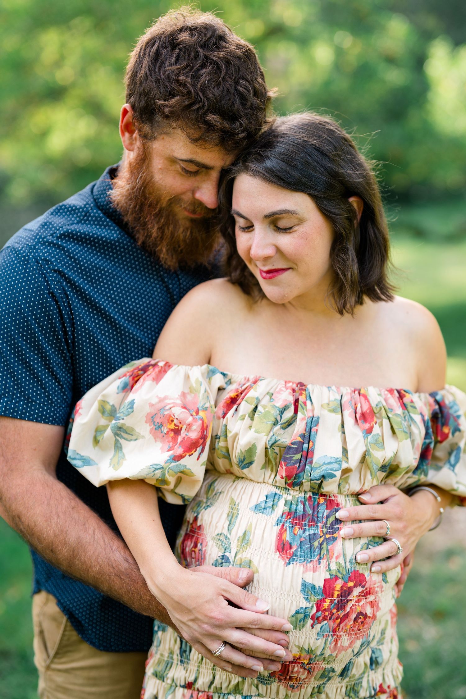Bearded man embracing pregnant woman in floral off-shoulder dress, both looking down at her baby bump in a lush garden.
