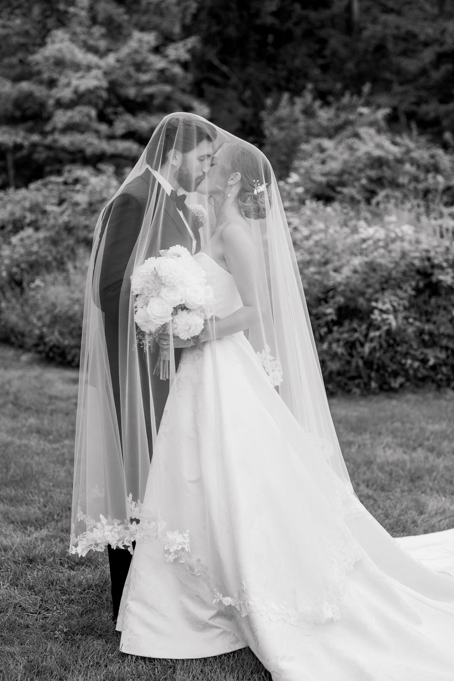 Black and white photo of bride and groom kissing under flowing lace-trimmed cathedral veil in garden.