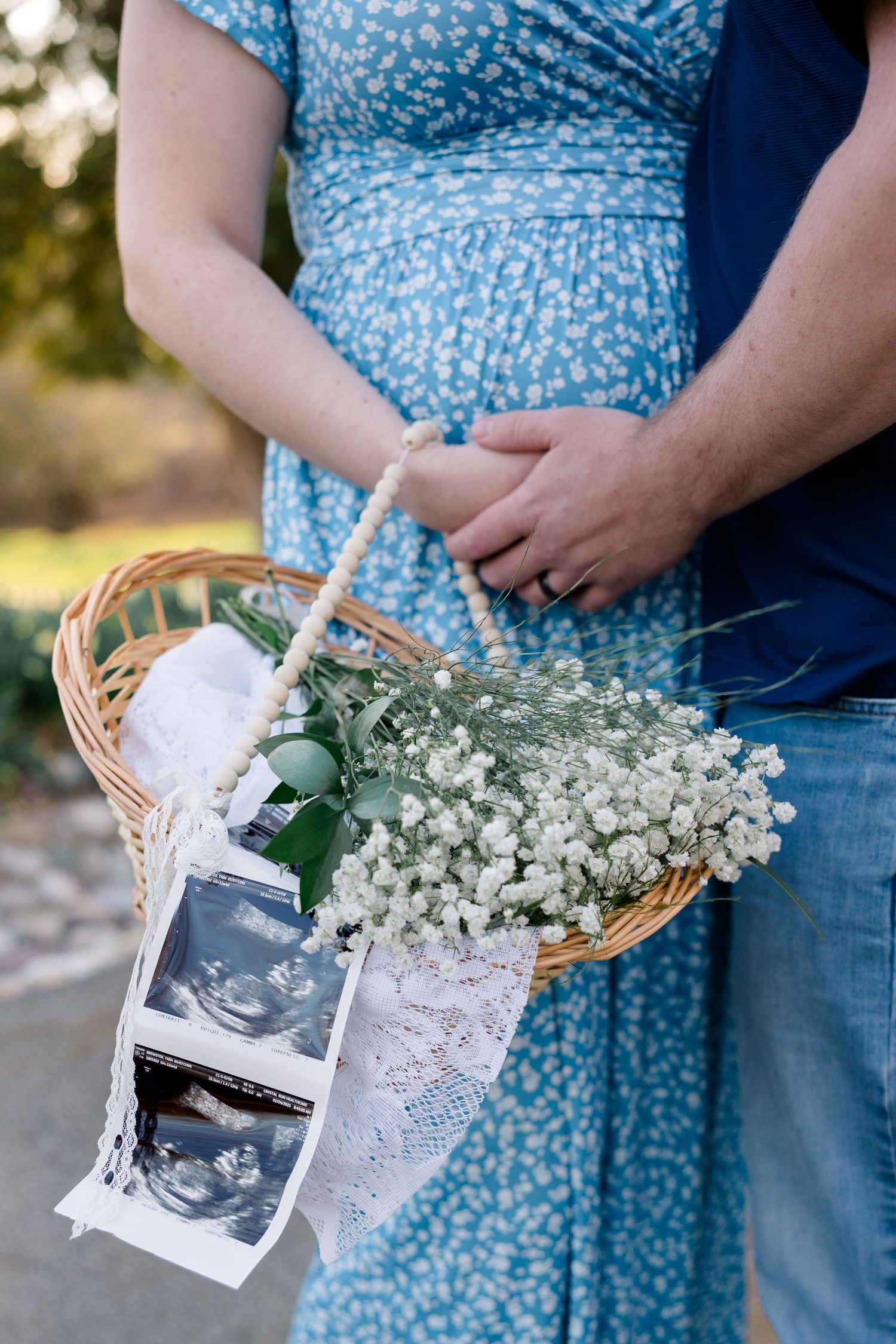 Someone in a blue floral dress holds a basket with baby's breath flowers and ultrasound photos adorned with lace.