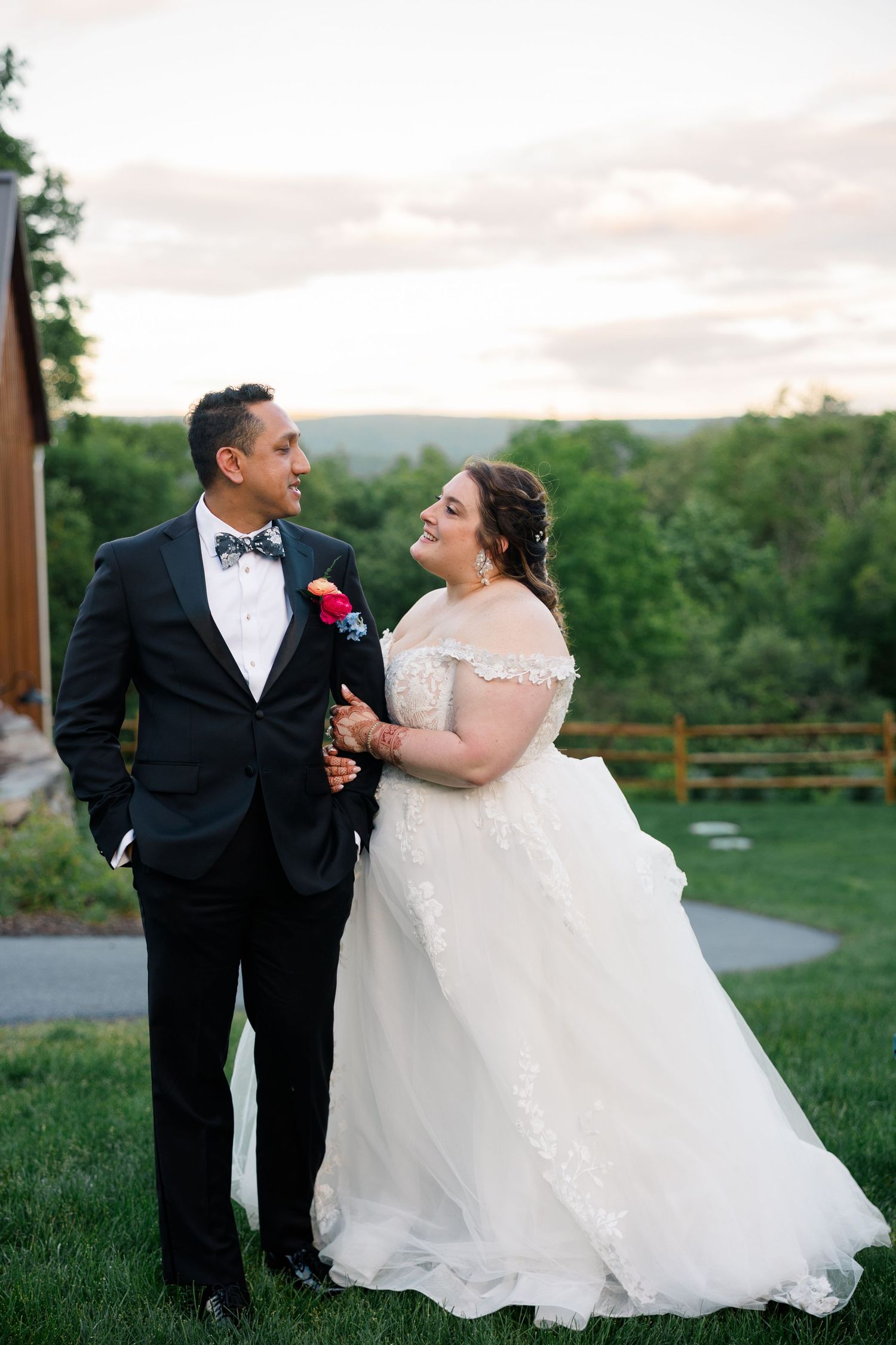 Elegant outdoor wedding scene with couple in formal attire posing on grassy hillside at sunset.