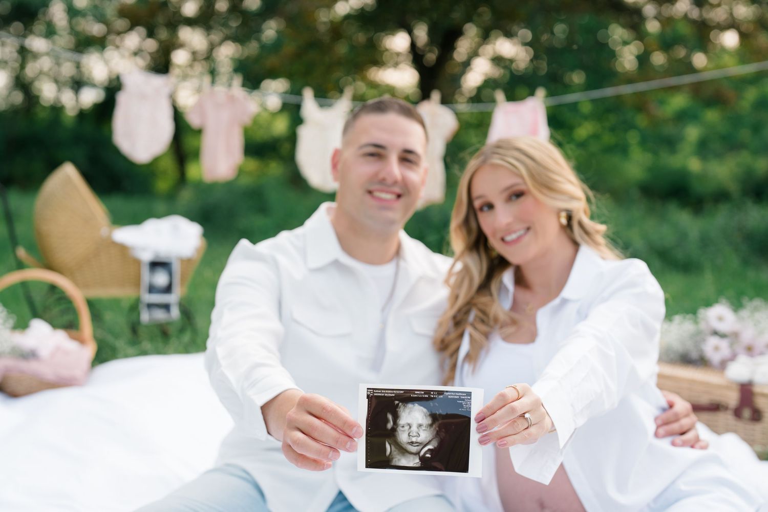 Happy couple in white outfits sit outdoors holding ultrasound photo with baby clothes on clothesline in background.