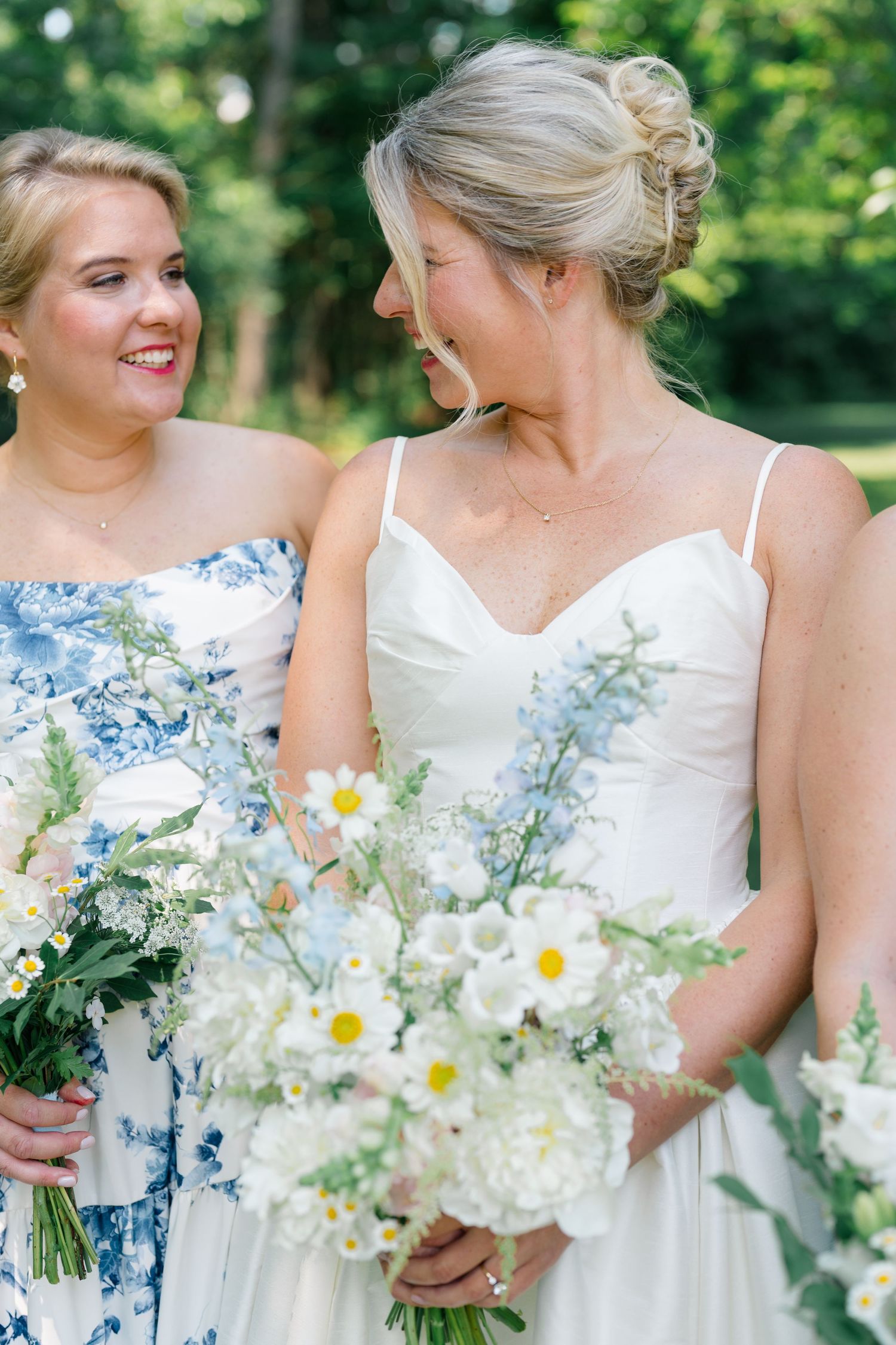 Bride in white gown and bridesmaid in blue floral dress share a joyful moment holding white daisy bouquets.