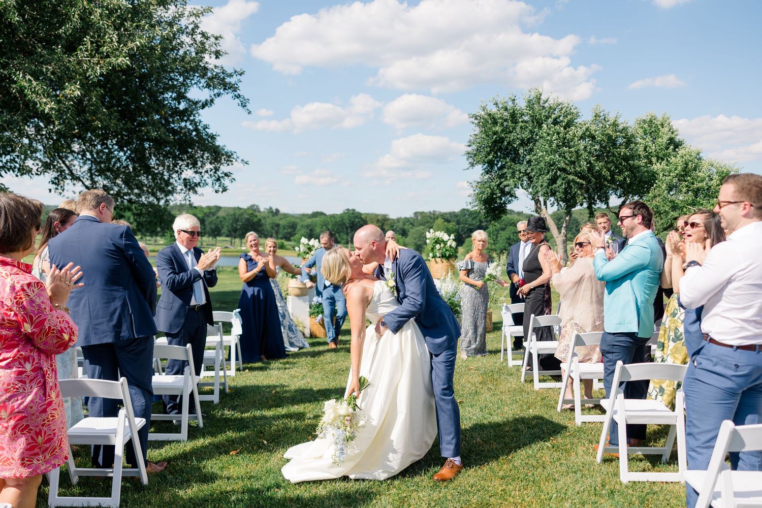 Bride and groom share a kiss at outdoor wedding ceremony while guests applaud along white chair-lined aisle.