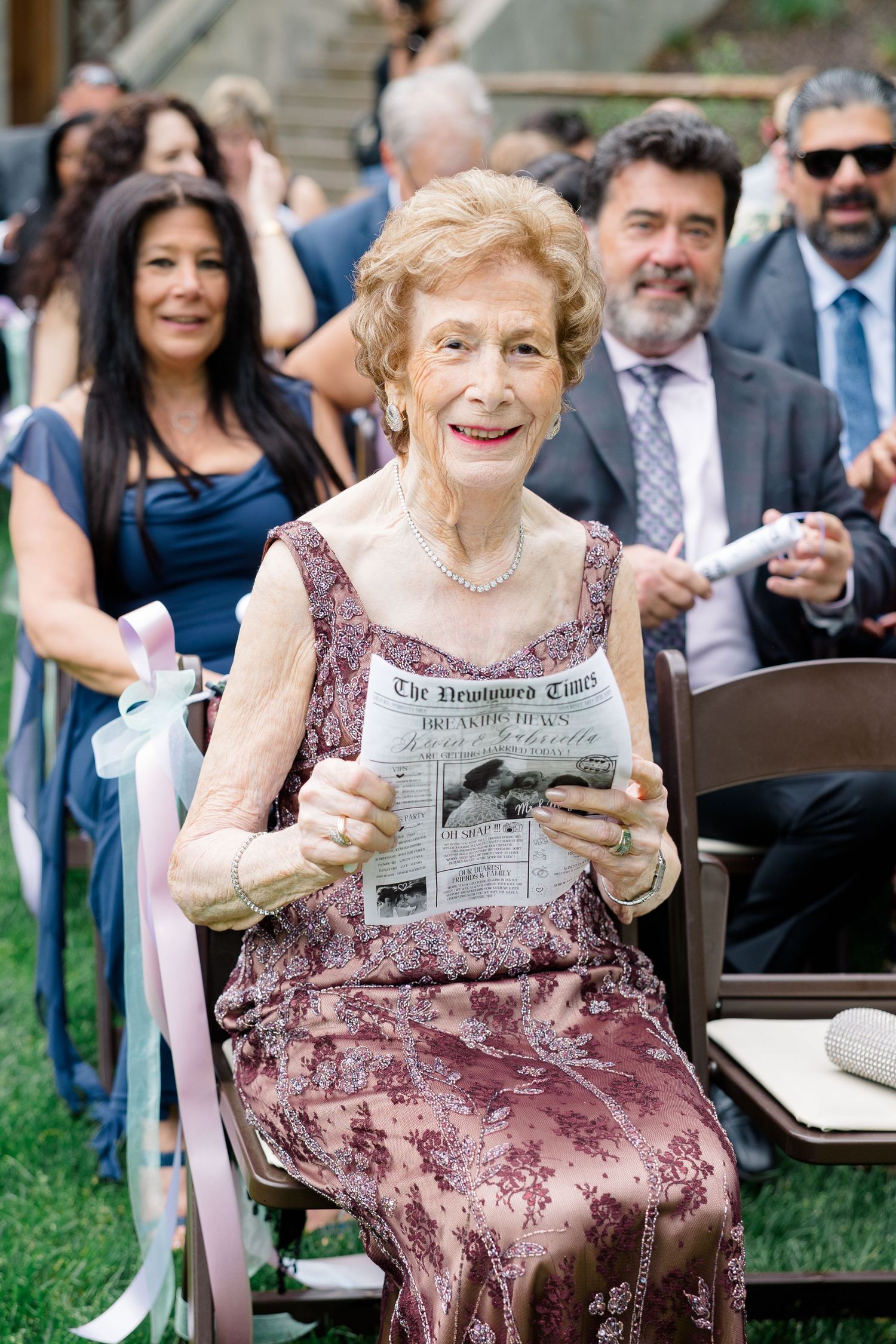 An elegant senior woman in a mauve gown reads a newspaper while sitting outdoors at an event.