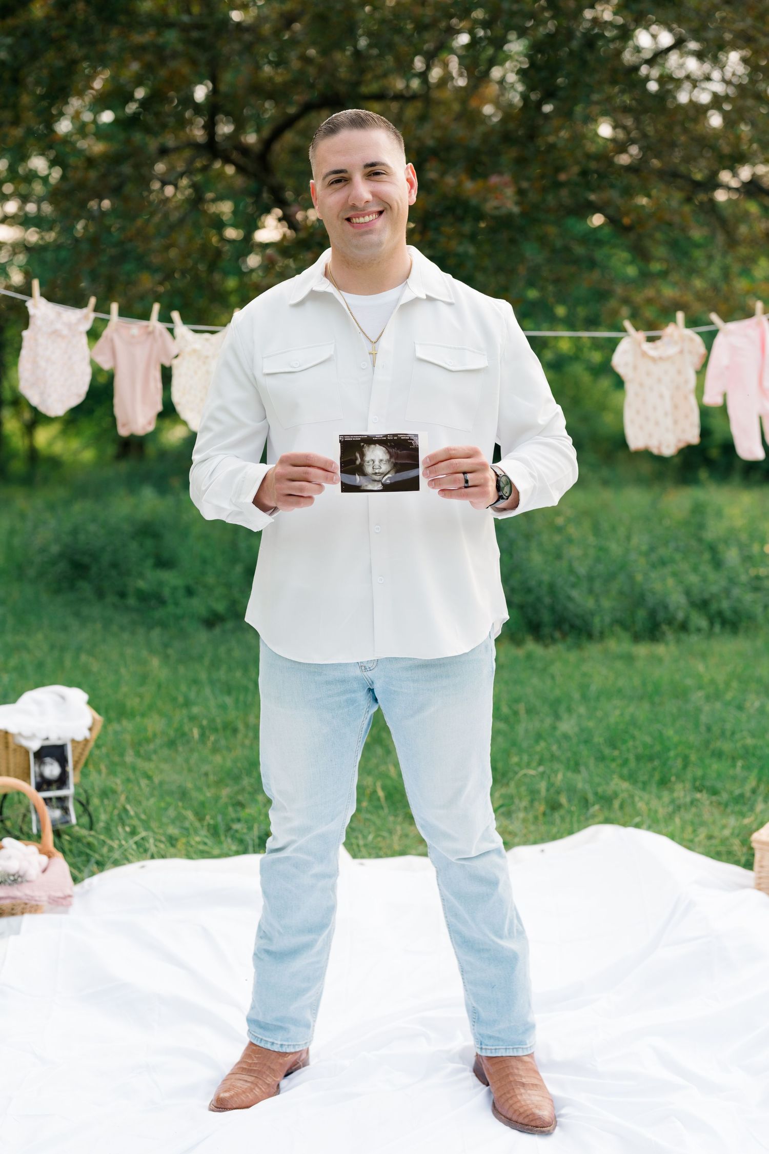 Person in white shirt and light jeans stands on white blanket showing ultrasound photos with baby items hanging.