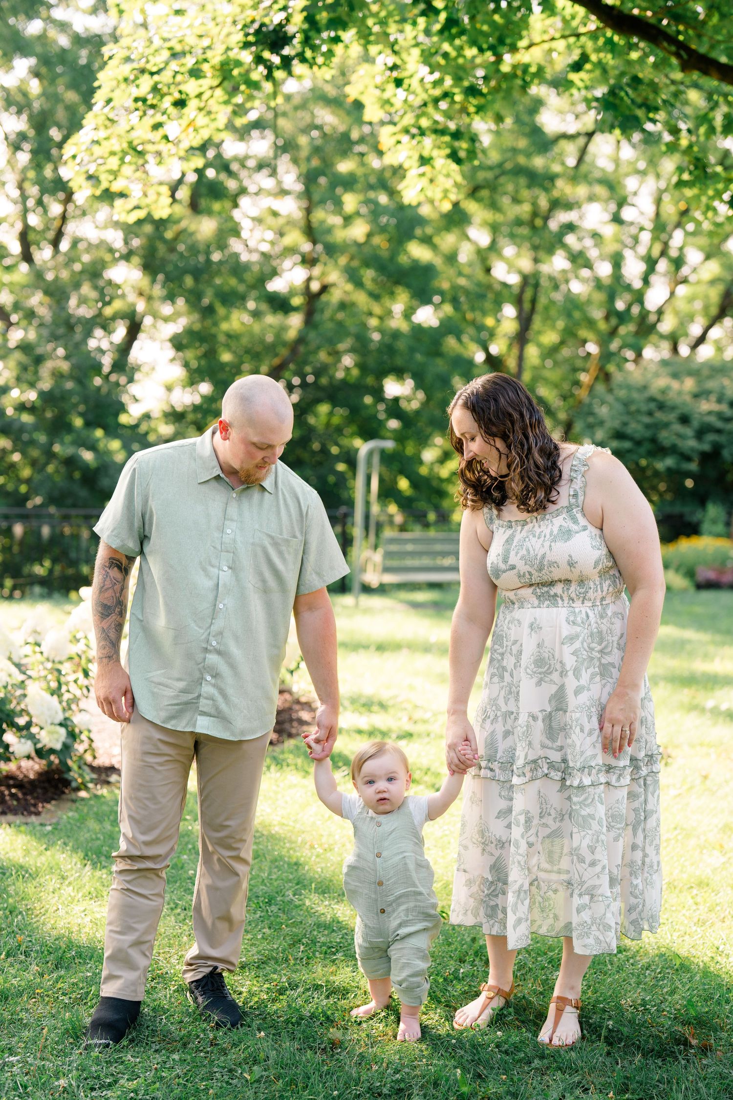 Parents help their baby take first steps on green grass during a sunny outdoor family photo session.
