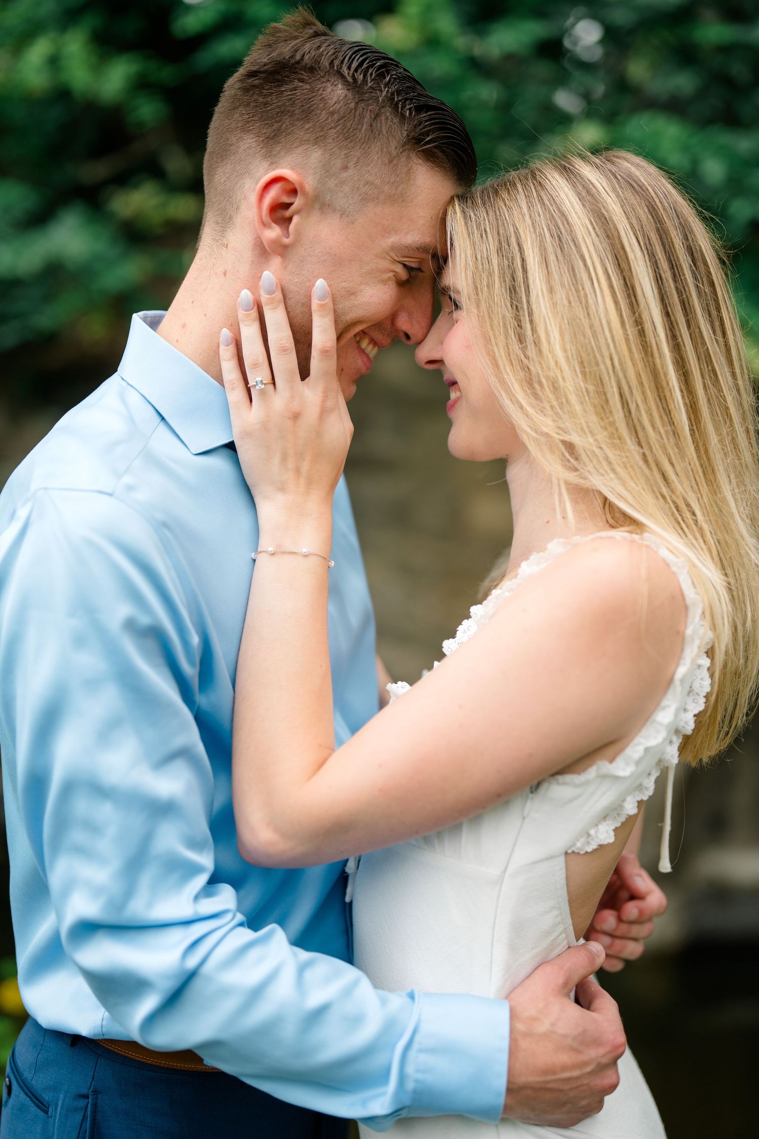 A smiling couple in love almost kissing outdoors, woman in white dress showing engagement ring, man in blue shirt embracing her.