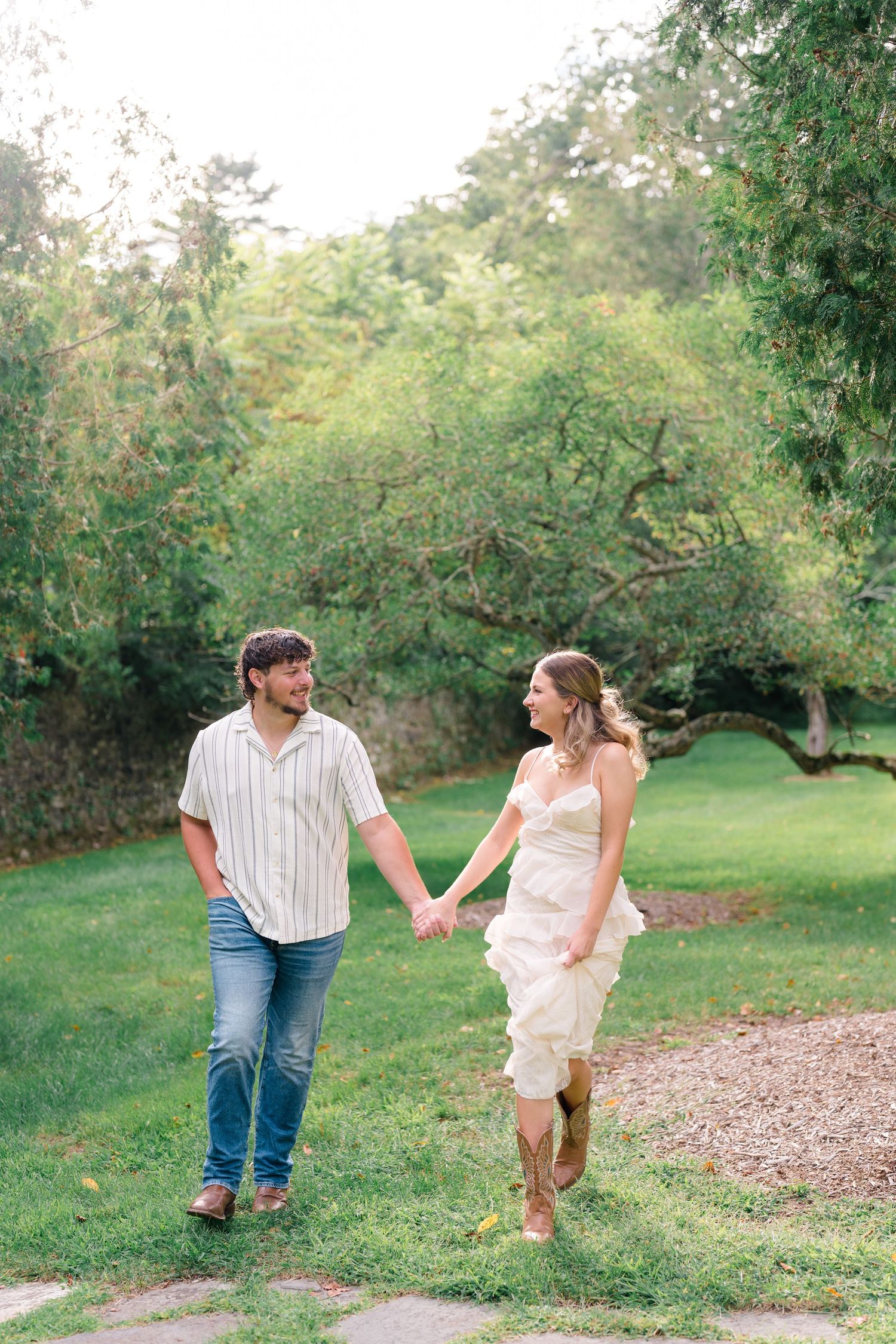 Couple holding hands and walking through lush green park during casual outdoor engagement photo session.