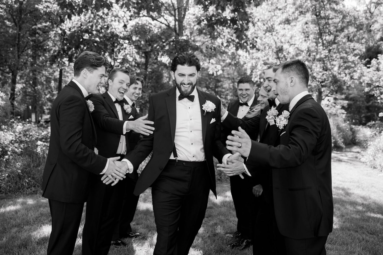 Groom laughing with groomsmen in black tuxedos outdoors in a candid black and white wedding party photo.