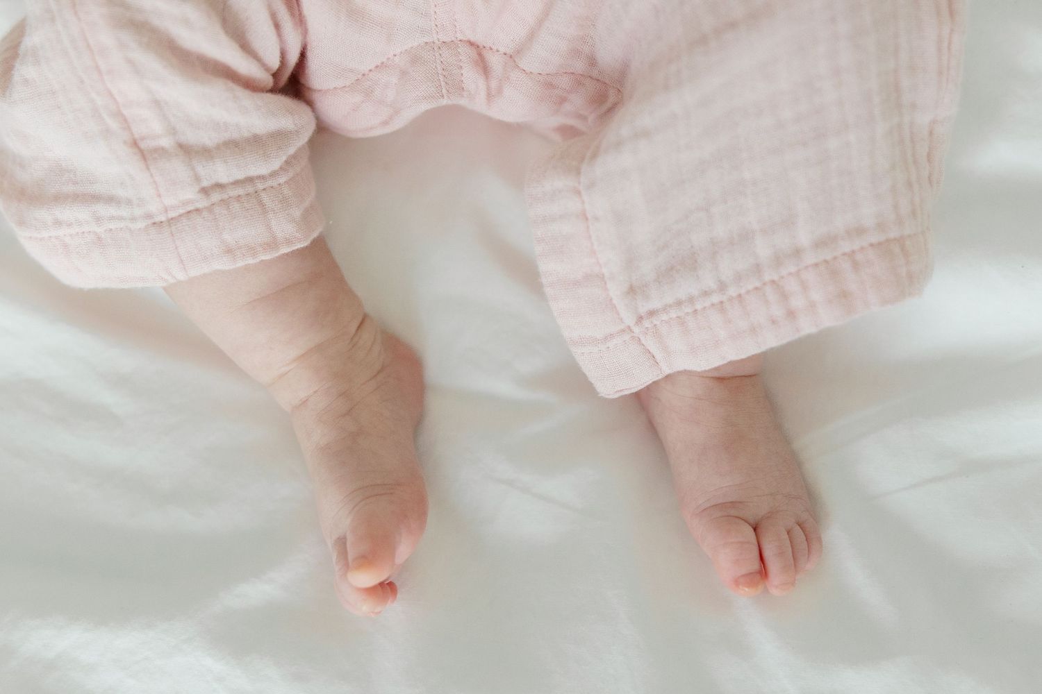 A baby wearing pink striped pajamas plays with their feet while lying on white bedding.