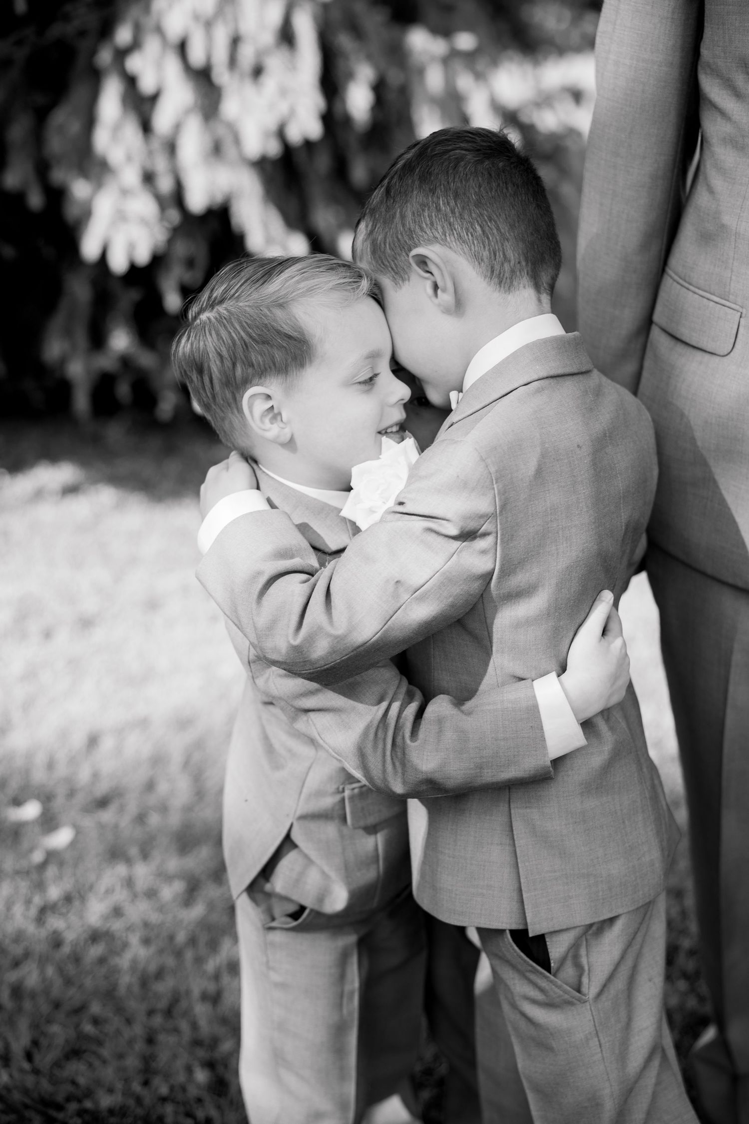 Two young boys in gray suits share a touching brotherly embrace outdoors in black and white photography.