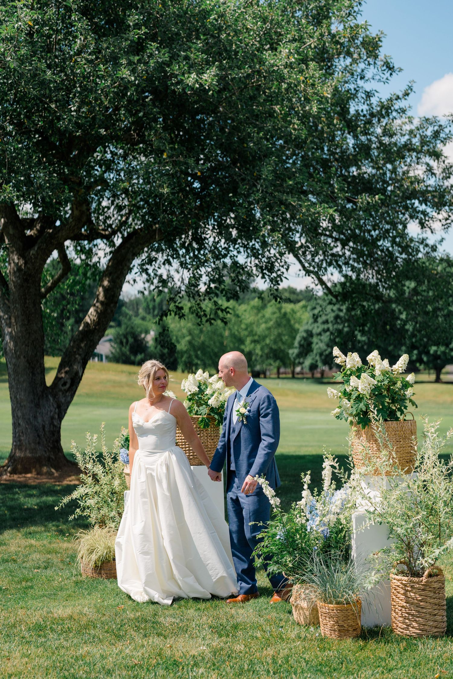 Bride and groom walk together at outdoor ceremony altar decorated with white florals and wicker baskets.