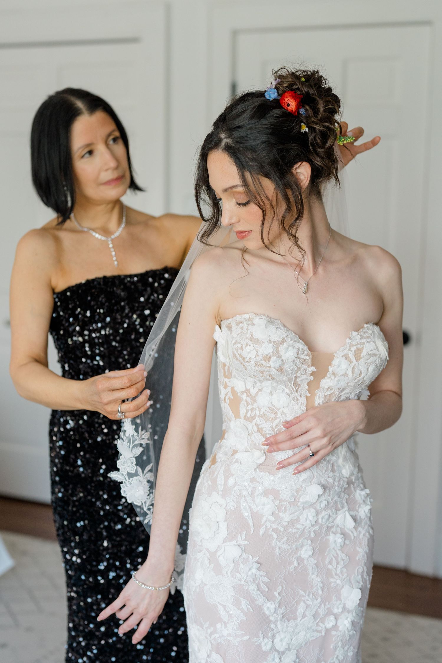 A bride in a delicate lace gown getting help from a bridesmaid wearing a black sequin dress.