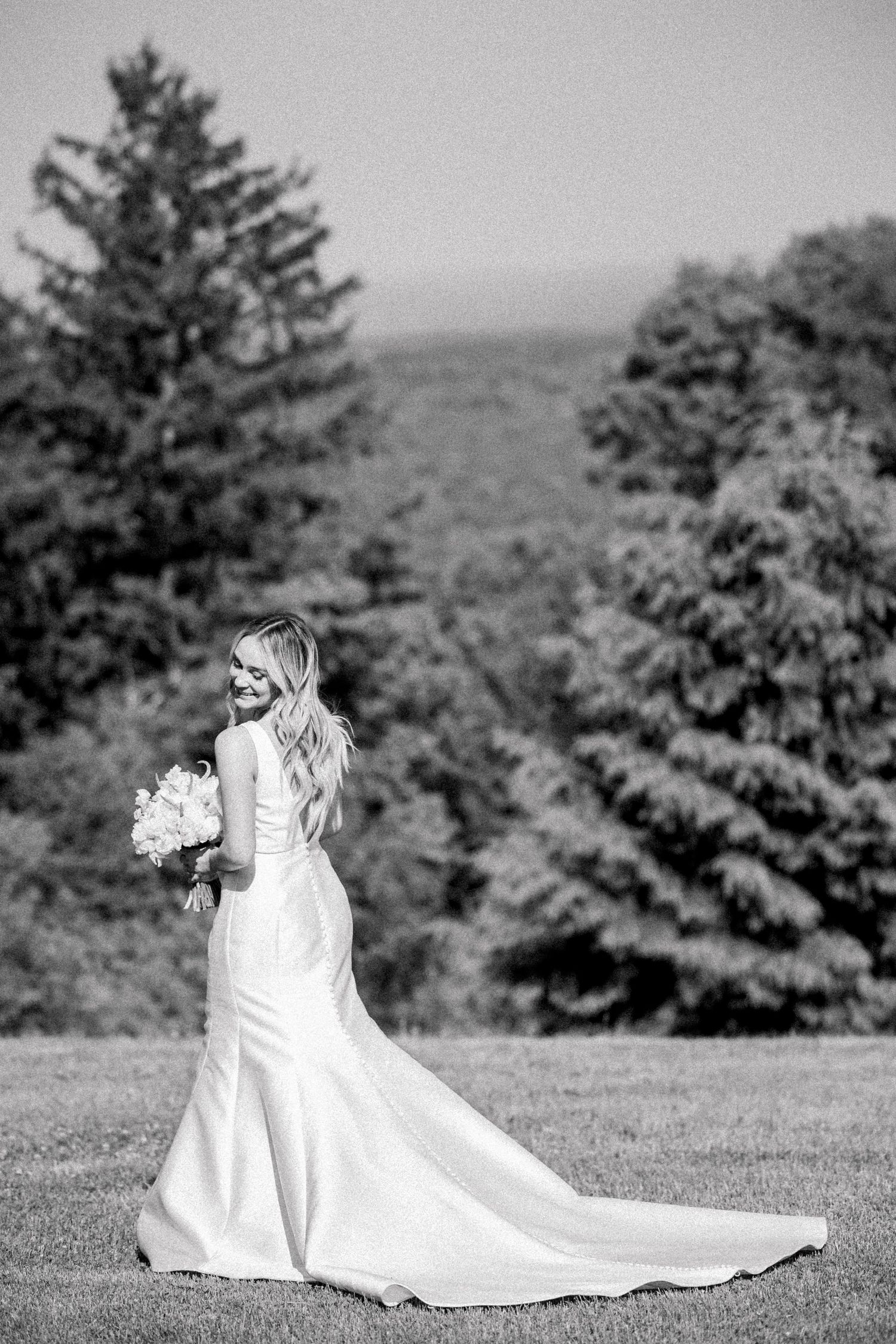 A bride in an elegant white wedding gown with a flowing train stands in a scenic outdoor setting among evergreen trees.