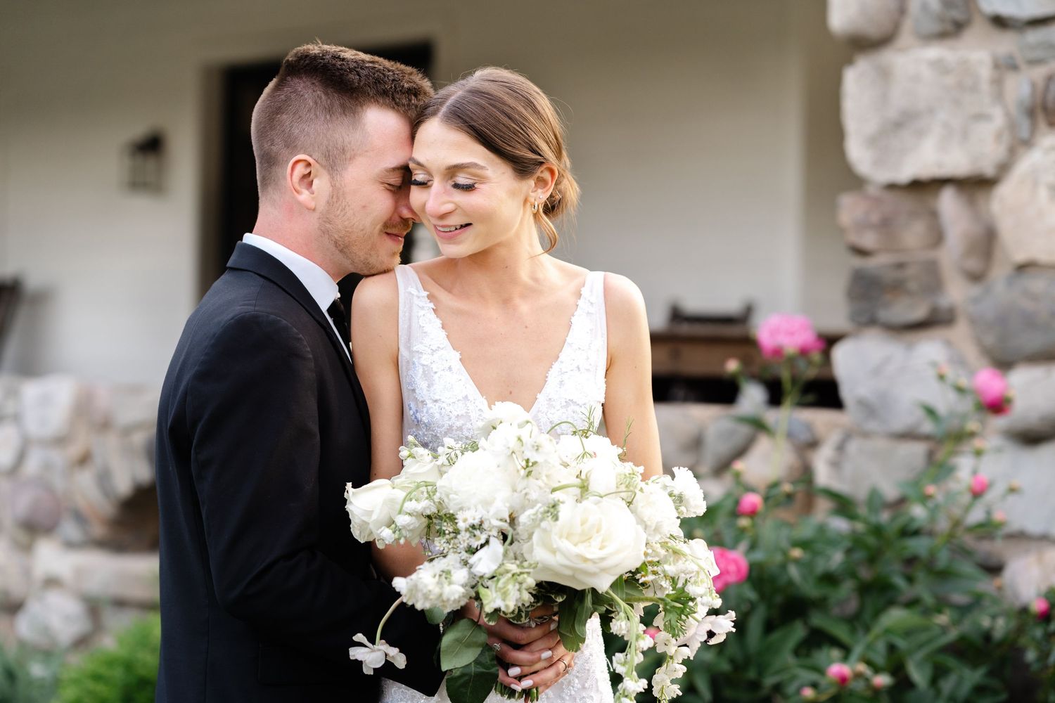 A wedding couple embraces outdoors near a stone wall with white floral bouquets in a romantic moment.