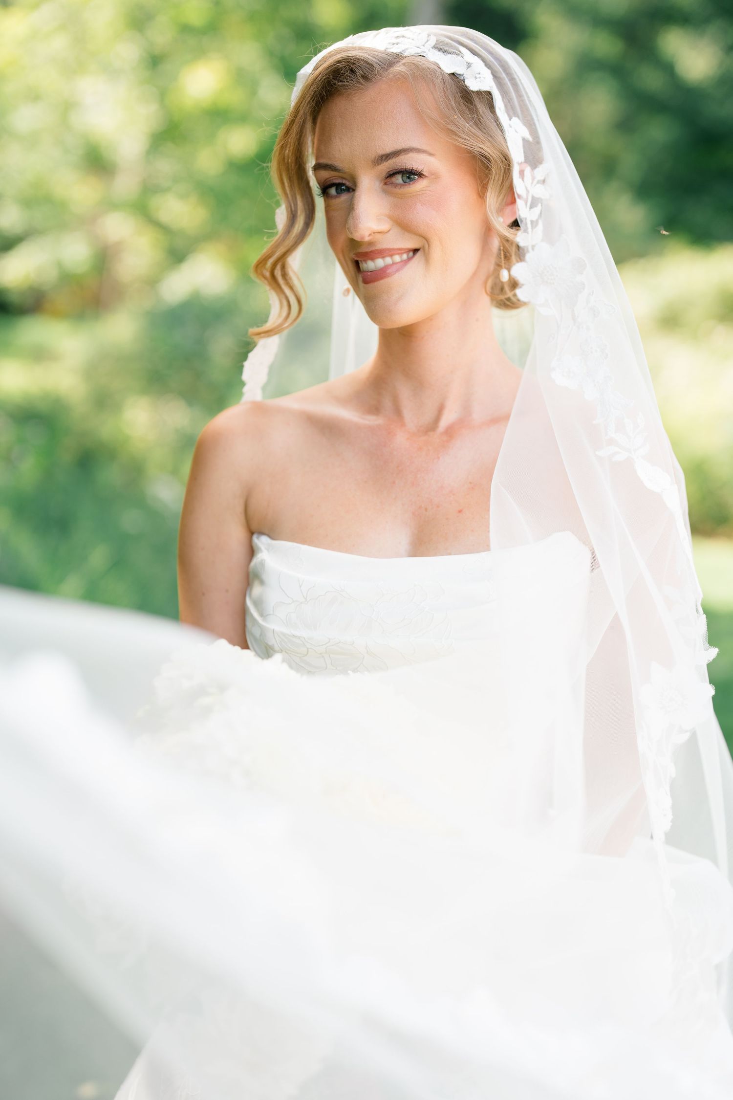 Smiling bride in white strapless wedding gown with lace veil flowing in breeze in outdoor garden setting.