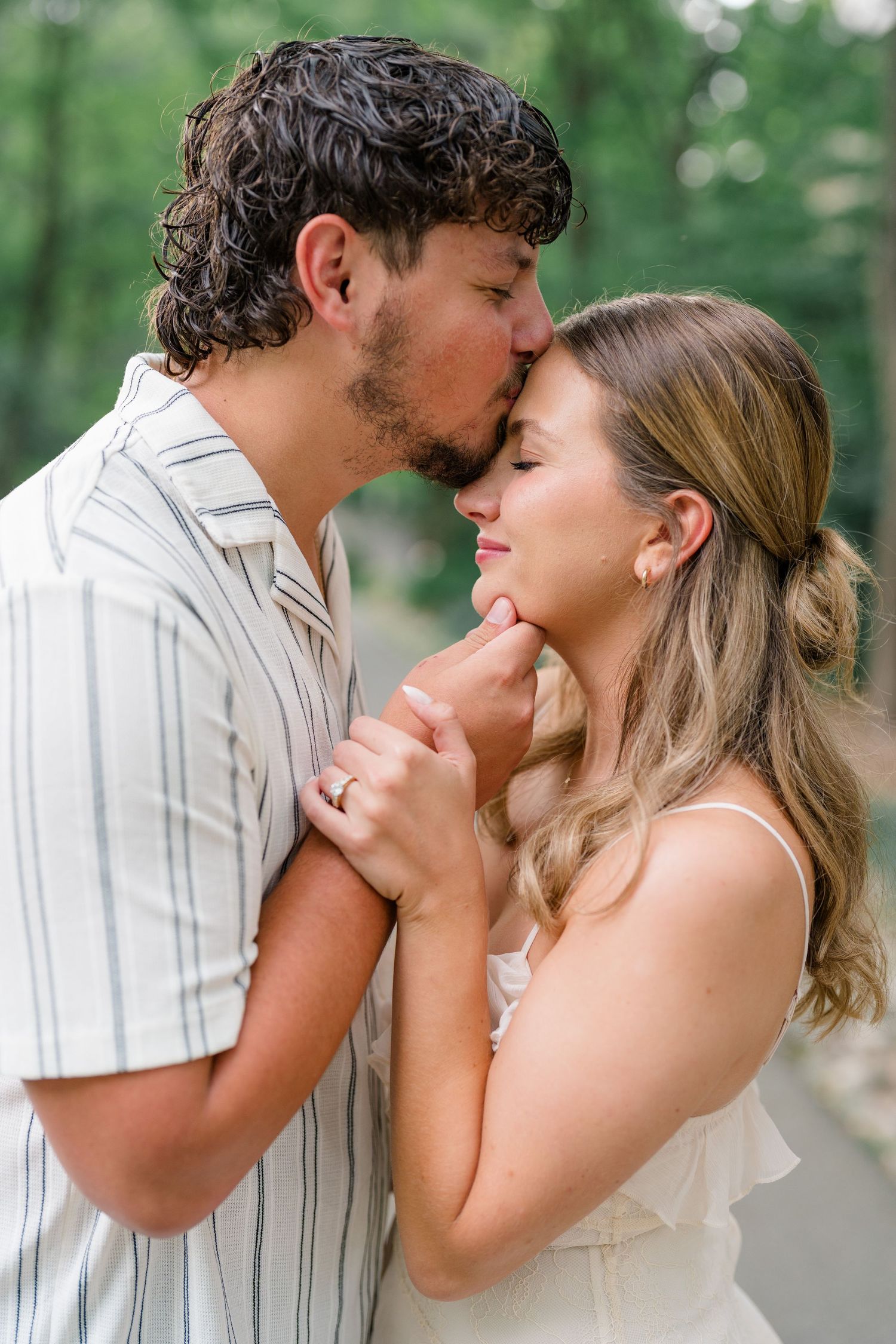 Man kissing woman on forehead while holding her face tenderly during romantic outdoor engagement photo session.