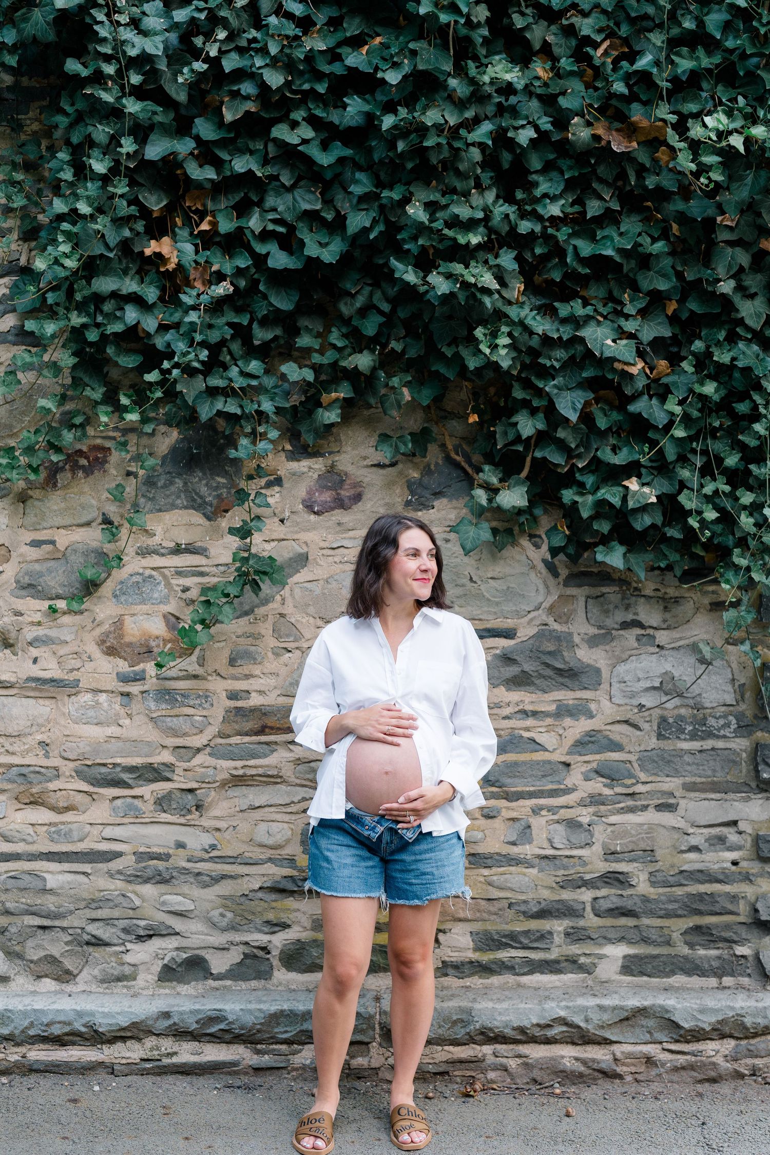 Pregnant woman in white shirt and denim shorts cradling baby bump against ivy-covered stone wall.