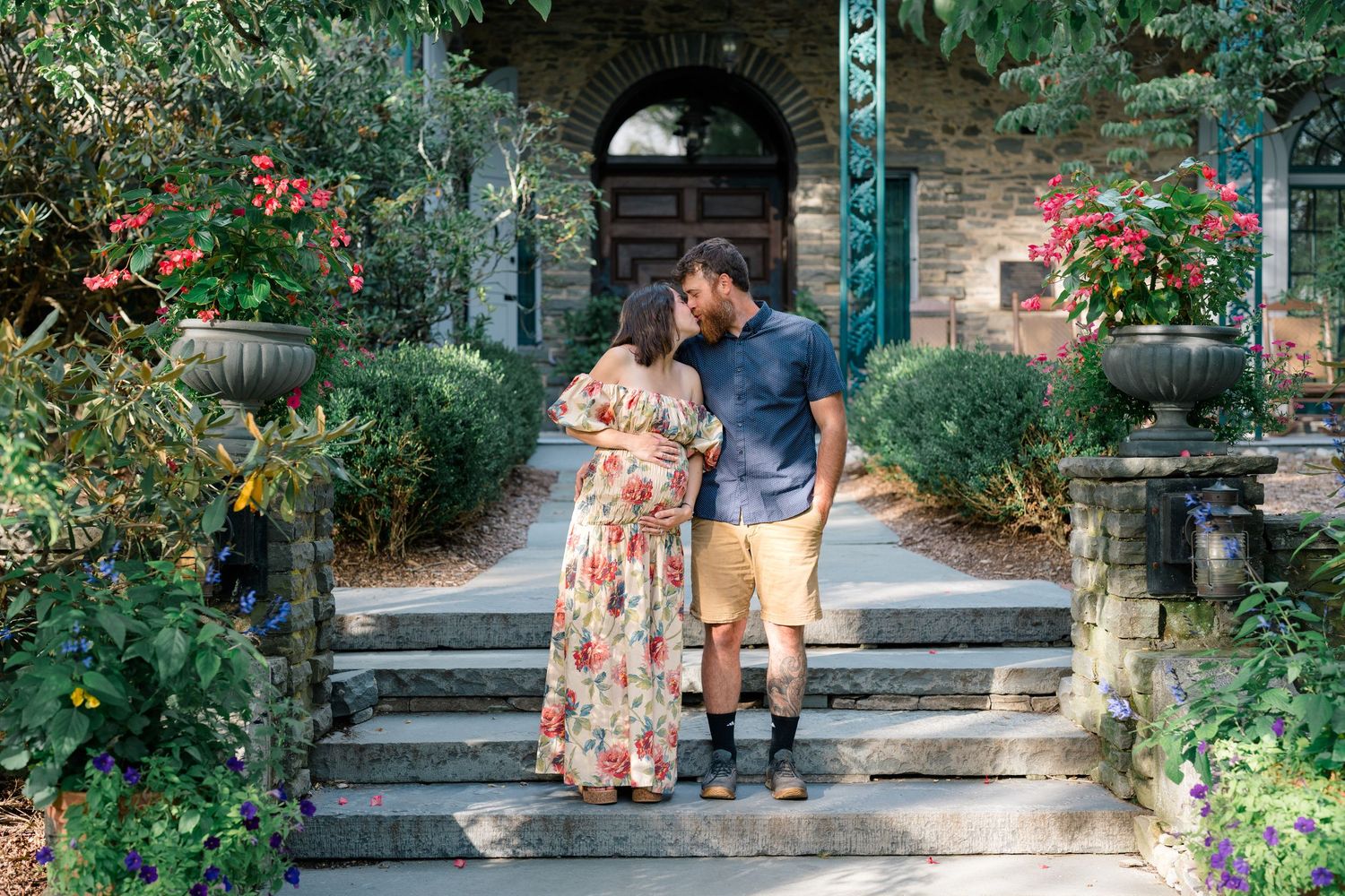 Pregnant couple sharing a kiss on garden steps surrounded by lush greenery and colorful flowers outside a stone building.