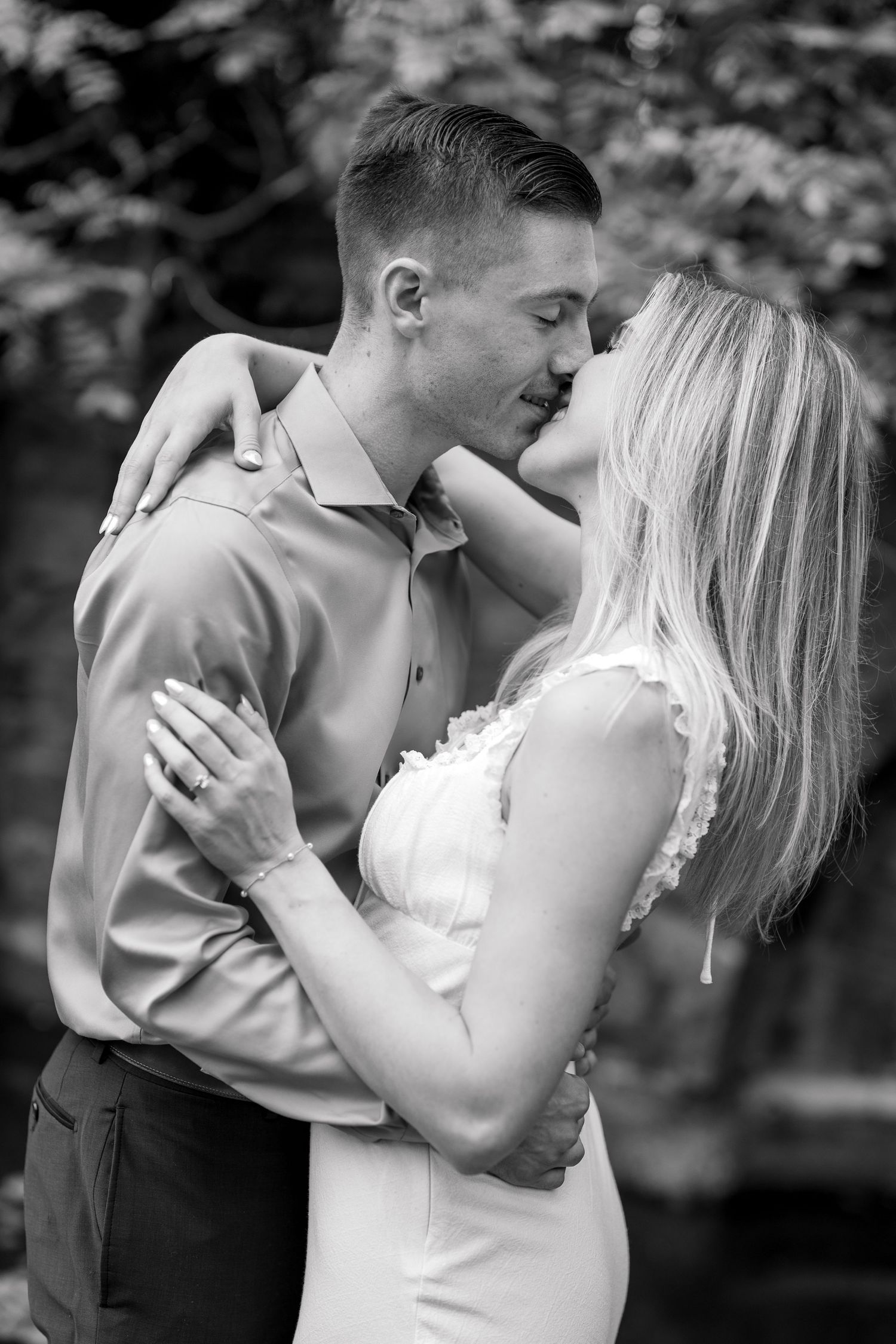 Black and white photo of a couple embracing and kissing outdoors, woman in white dress, man in button-up shirt.