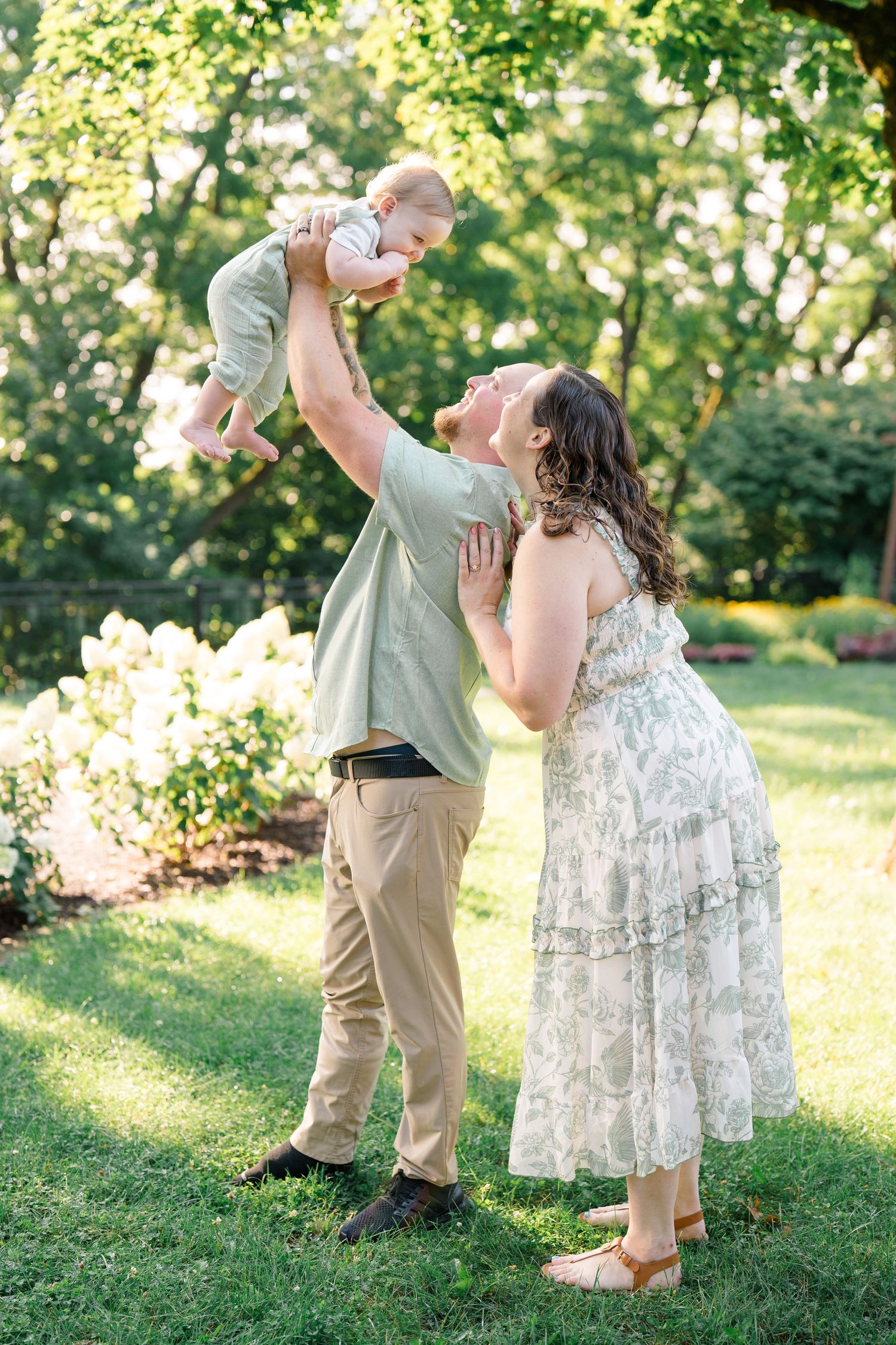 A joyful couple lifts their baby in a lush green garden, with the mother in a floral dress smiling warmly.